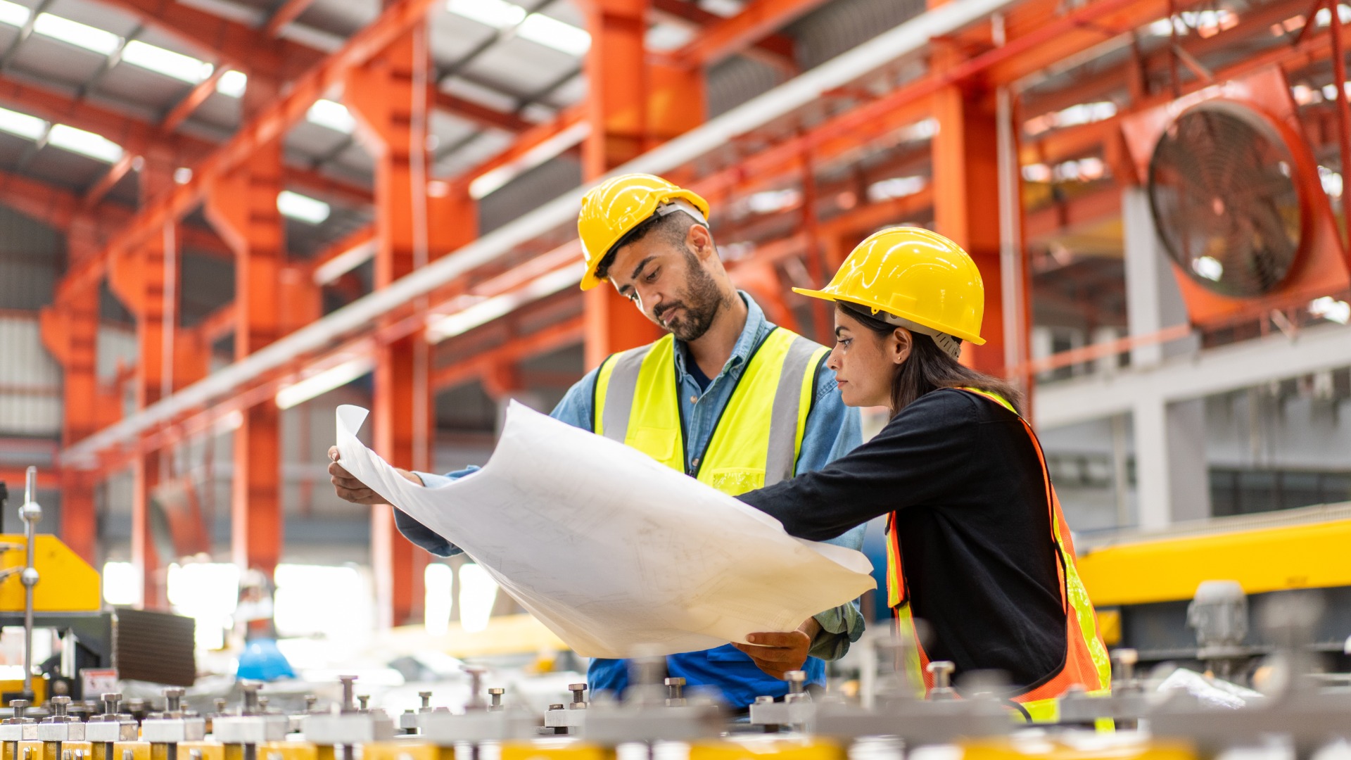A worker and an engineer in safety gear are reviewing information on a blueprint. They are in a large warehouse.