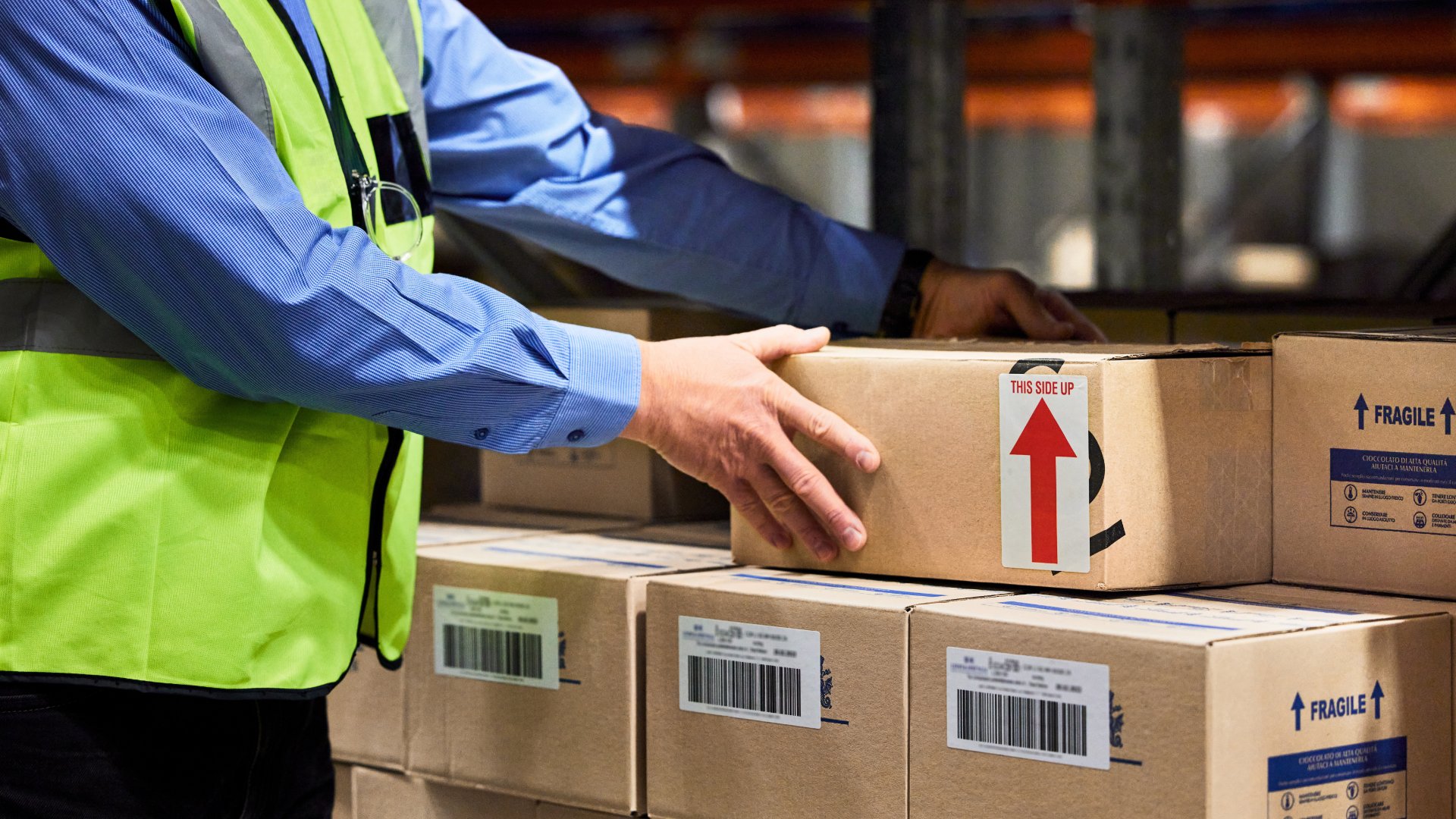 A man in a yellow safety vest is applying a shipping label to a cardboard box beside warehouse packing supplies.