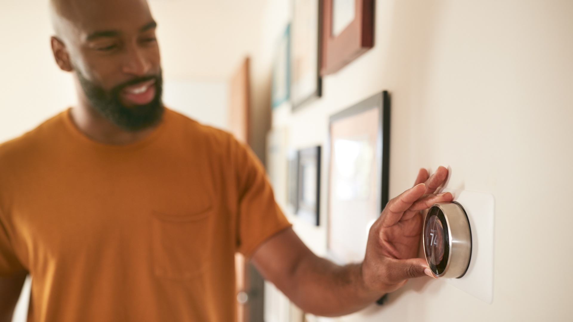 A man in a brown T-shirt adjusts a circular digital thermostat mounted on a white wall with picture frames.
