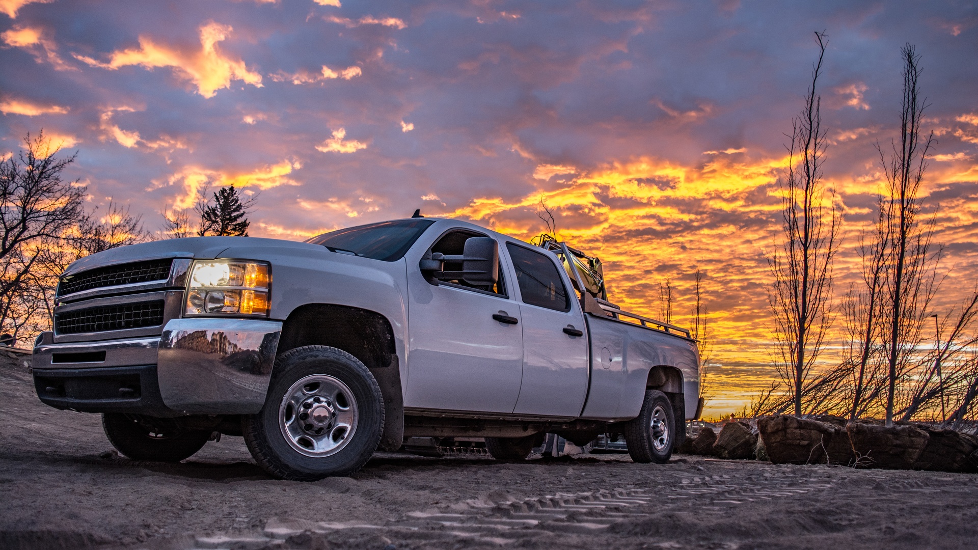 A truck parked outdoors. The headlights are on and the sun is low in the sky. There are bare trees around the truck.
