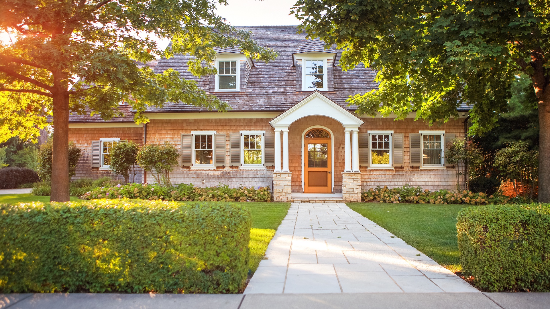 A classic brick house with an orange door and a concrete tile walkway. The house is surrounded by trees.