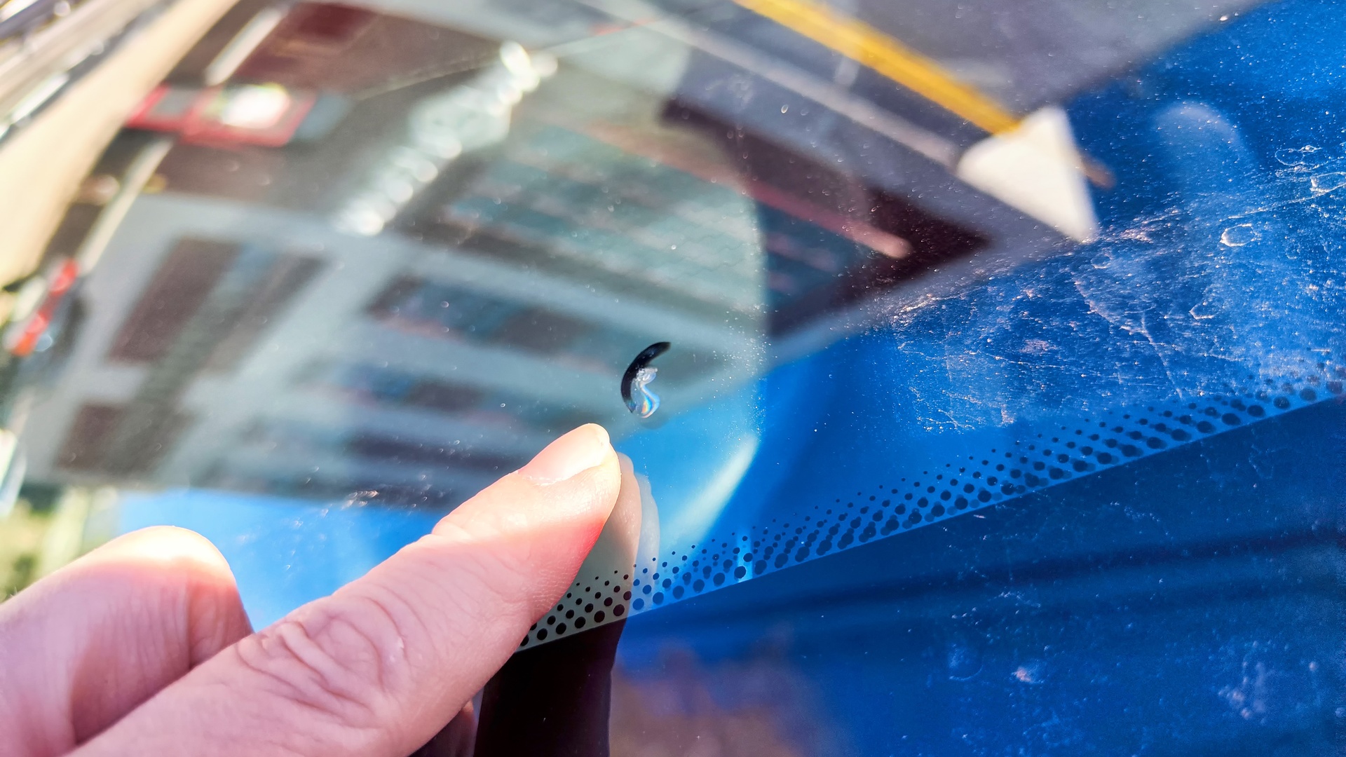 A close-up shot of someone's finger pointing to a small chip on a vehicle's windshield on a sunny day.
