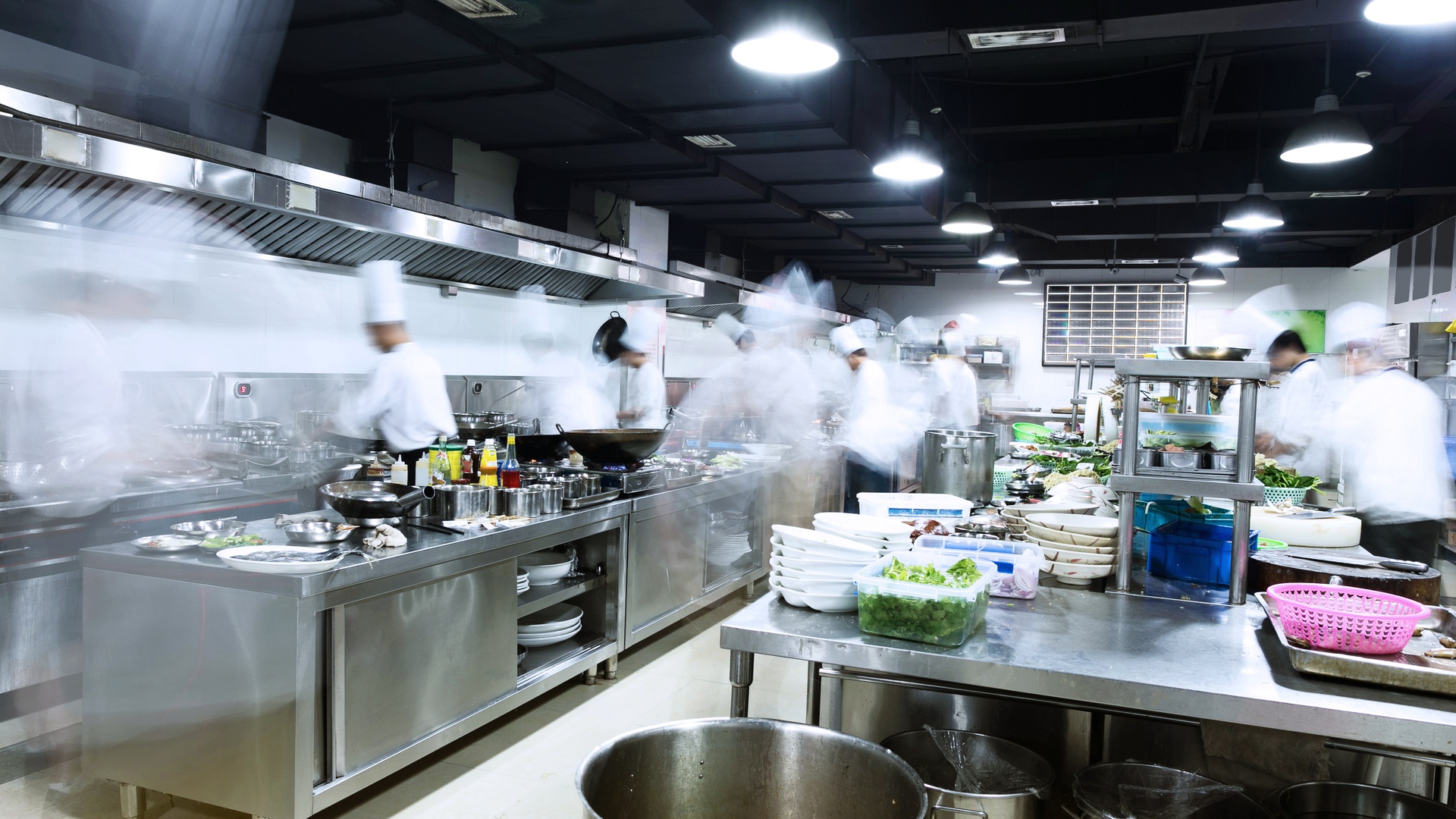 A large commercial kitchen with stainless-steel equipment, tables, pans, and baskets, with multiple chefs working.