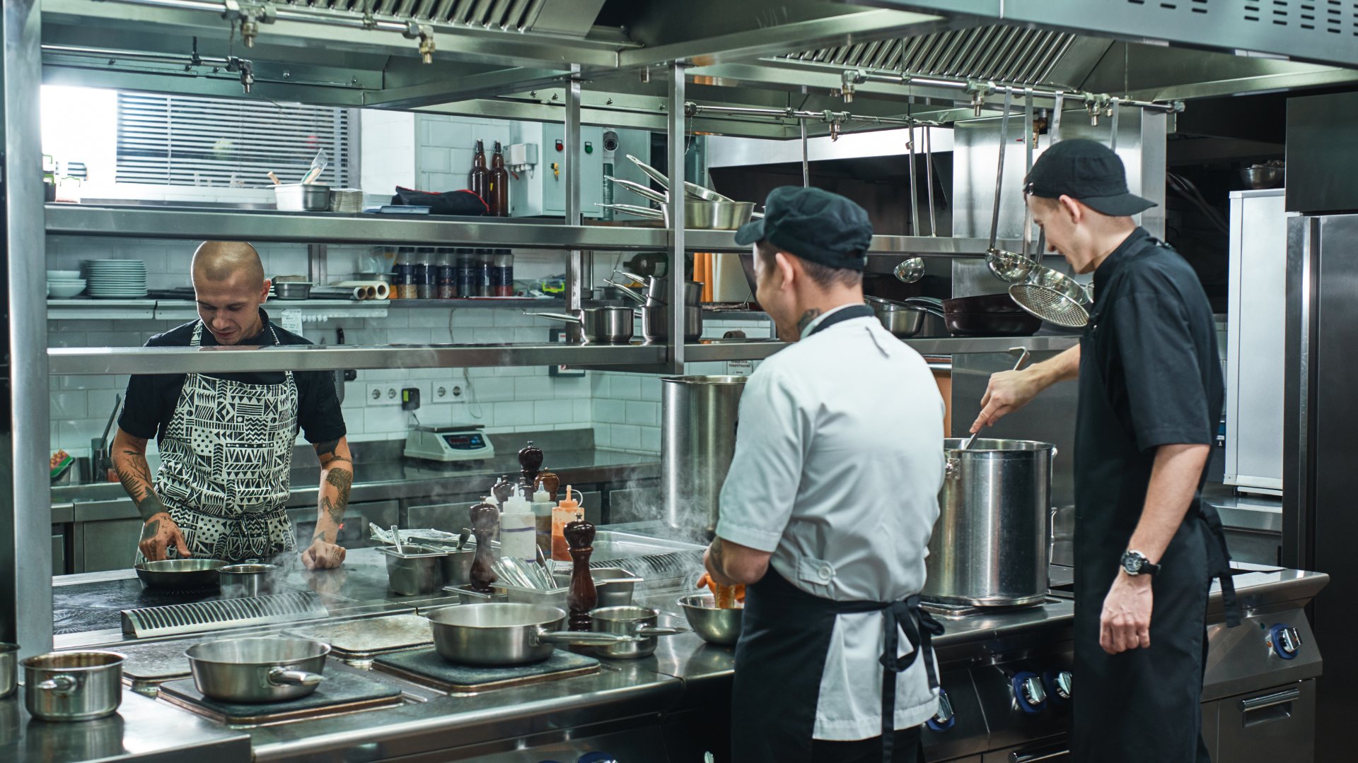 Three male workers wearing aprons cooking around a large stainless steel stove inside a commercial kitchen.