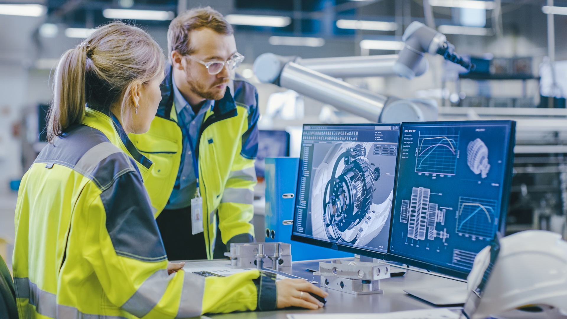 A man and a woman wearing protective yellow jackets, looking at two computer monitors inside a manufacturing warehouse.