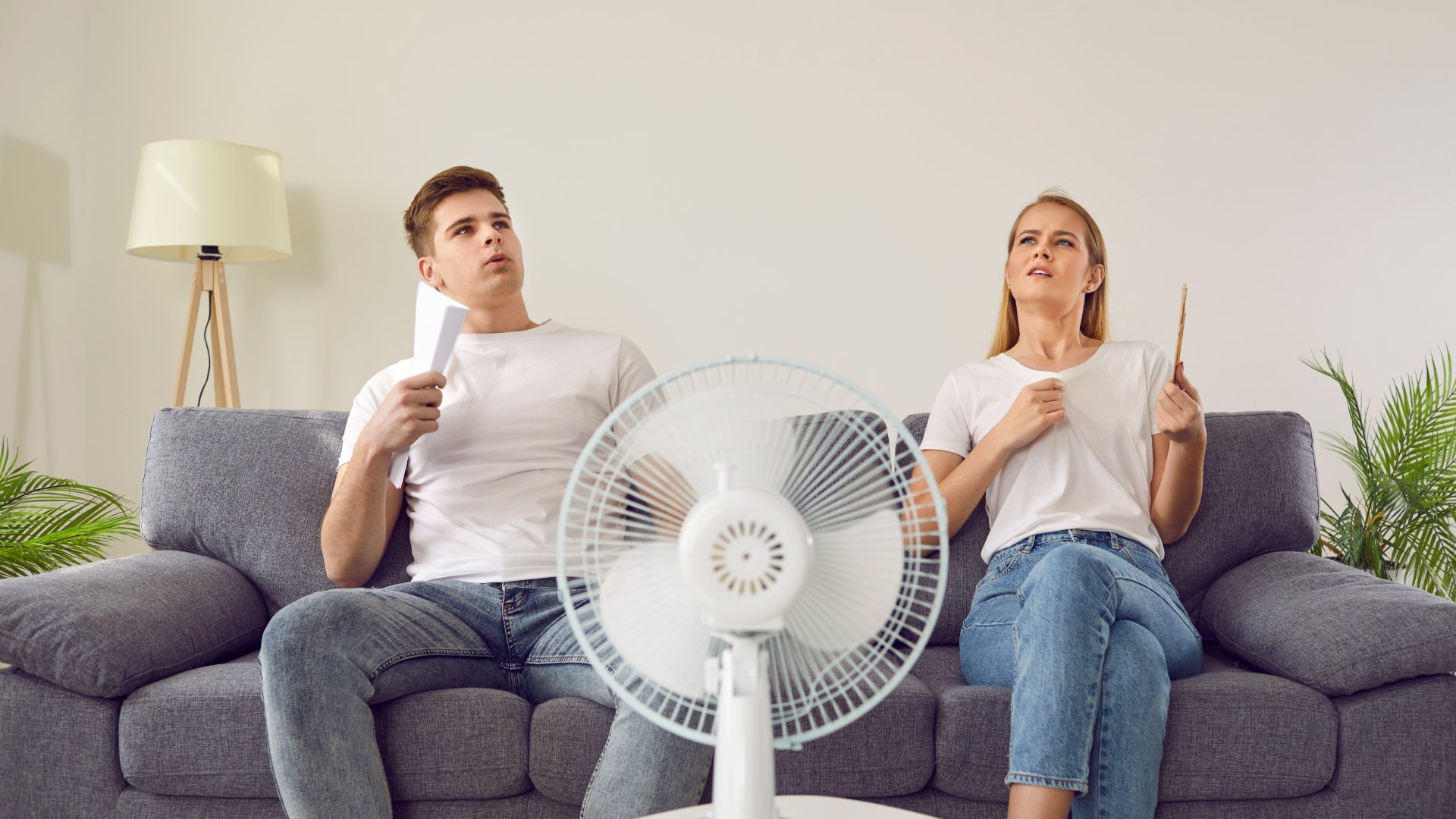A man and a woman sit on a couch in a room, acting as if they feel overheated. An electric fan blows on them.