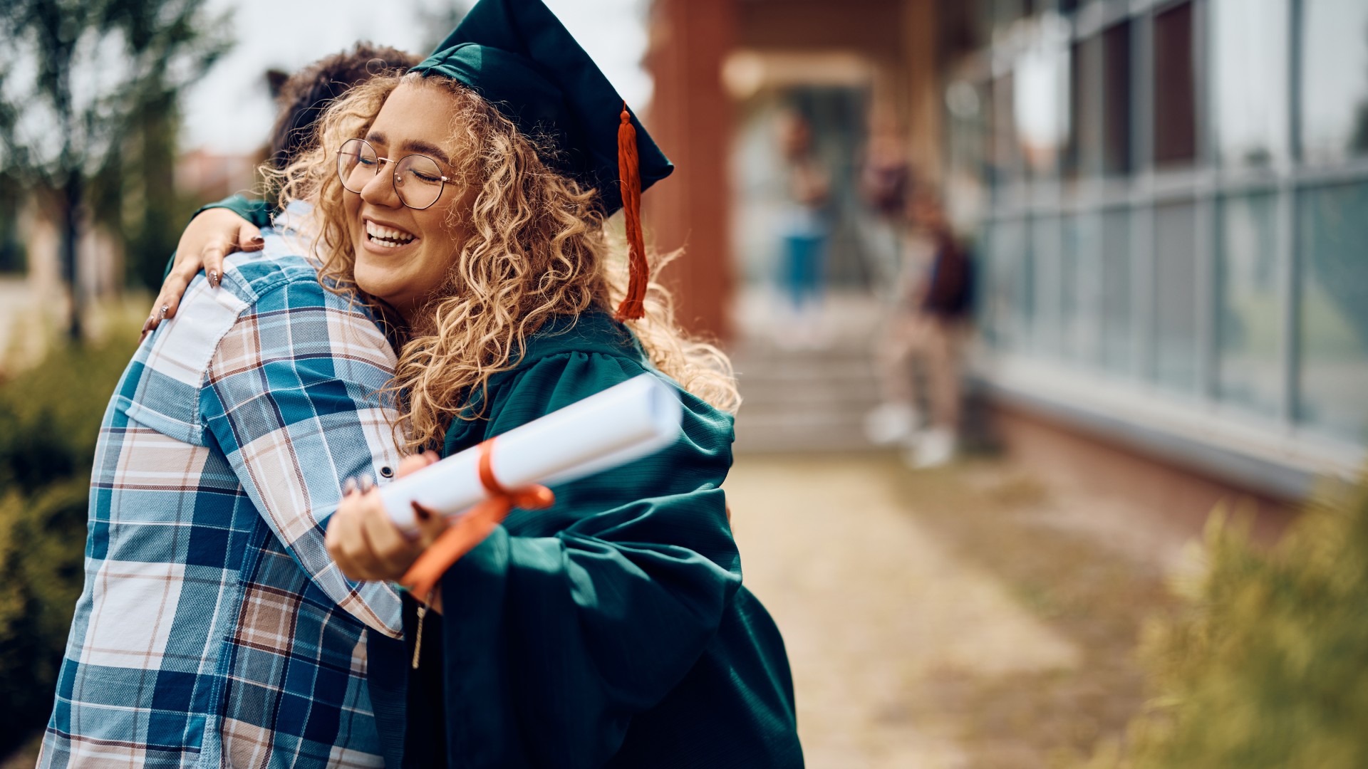 A womanwith curly hair and dressed in graduation attire is hugging a man as they are both outside a building.