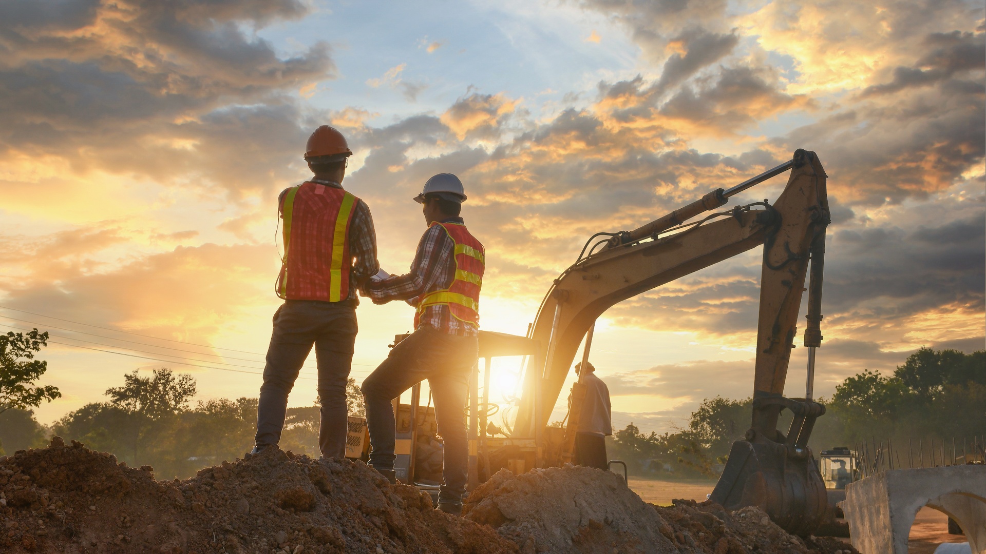 Construction engineers looking over a worksite from a tall mound of dirt. There is a excavator in front of them.