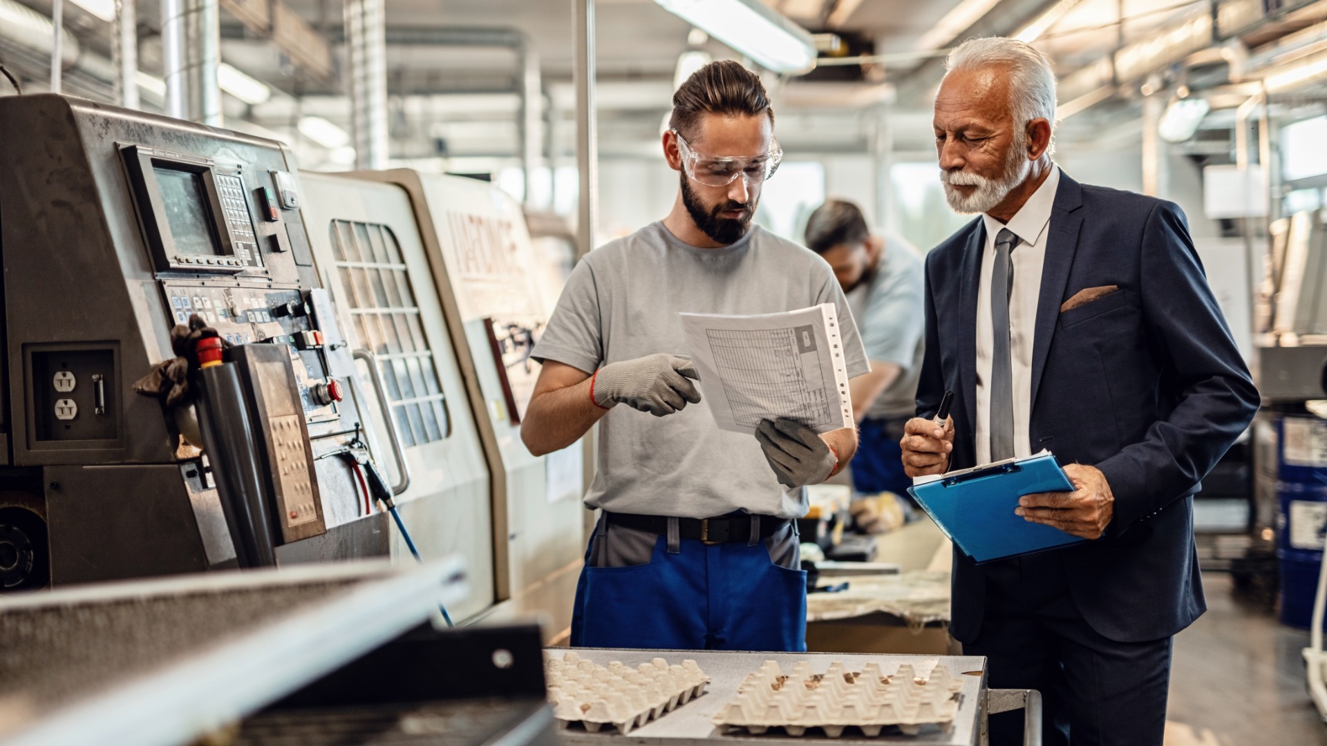 A manager and an employee look at a report while standing in an industrial facility. Machines surround the two men.