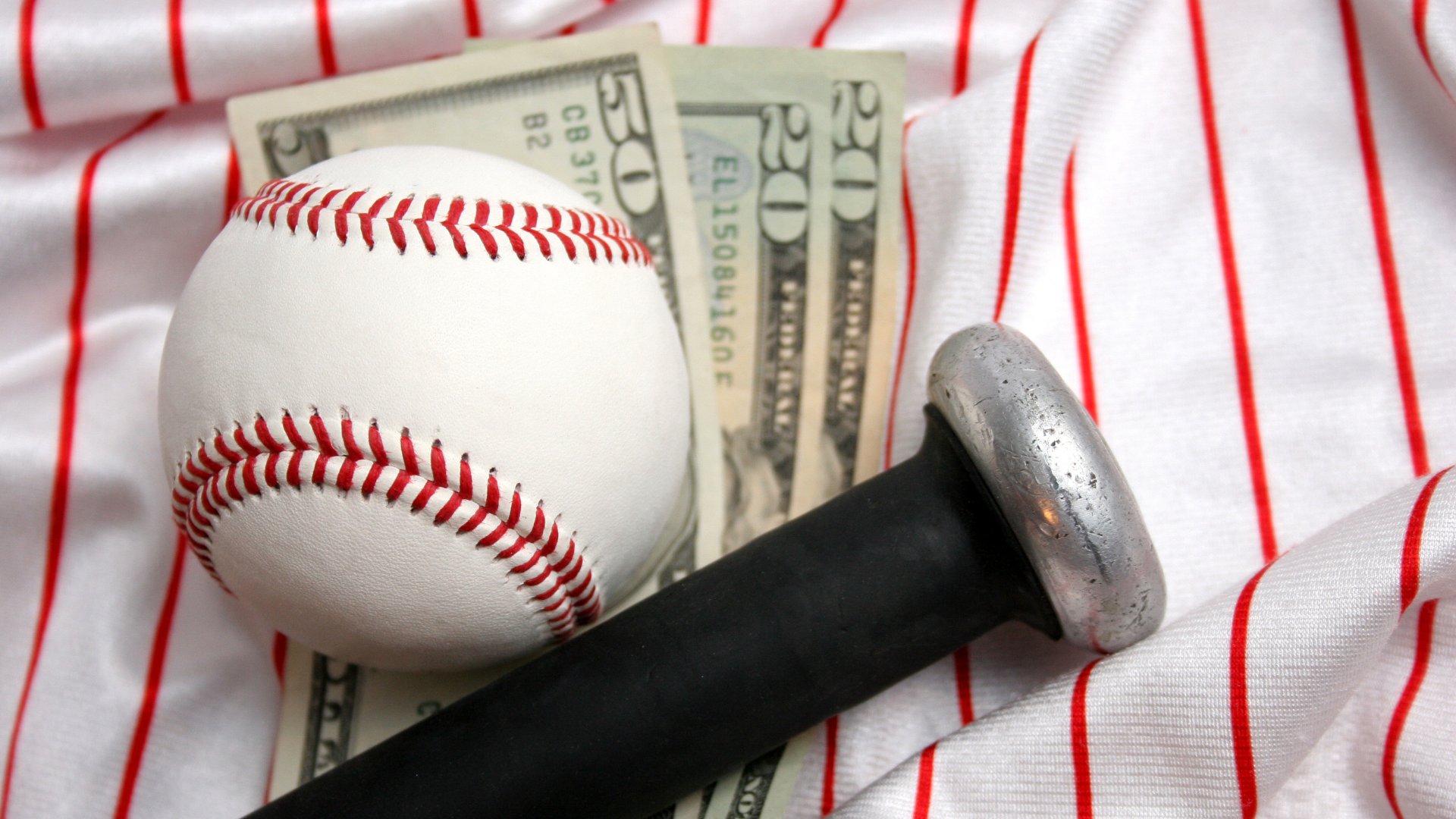 A close-up of a baseball and the knob of a baseball bat resting on three dollar bills and a white baseball jersey.