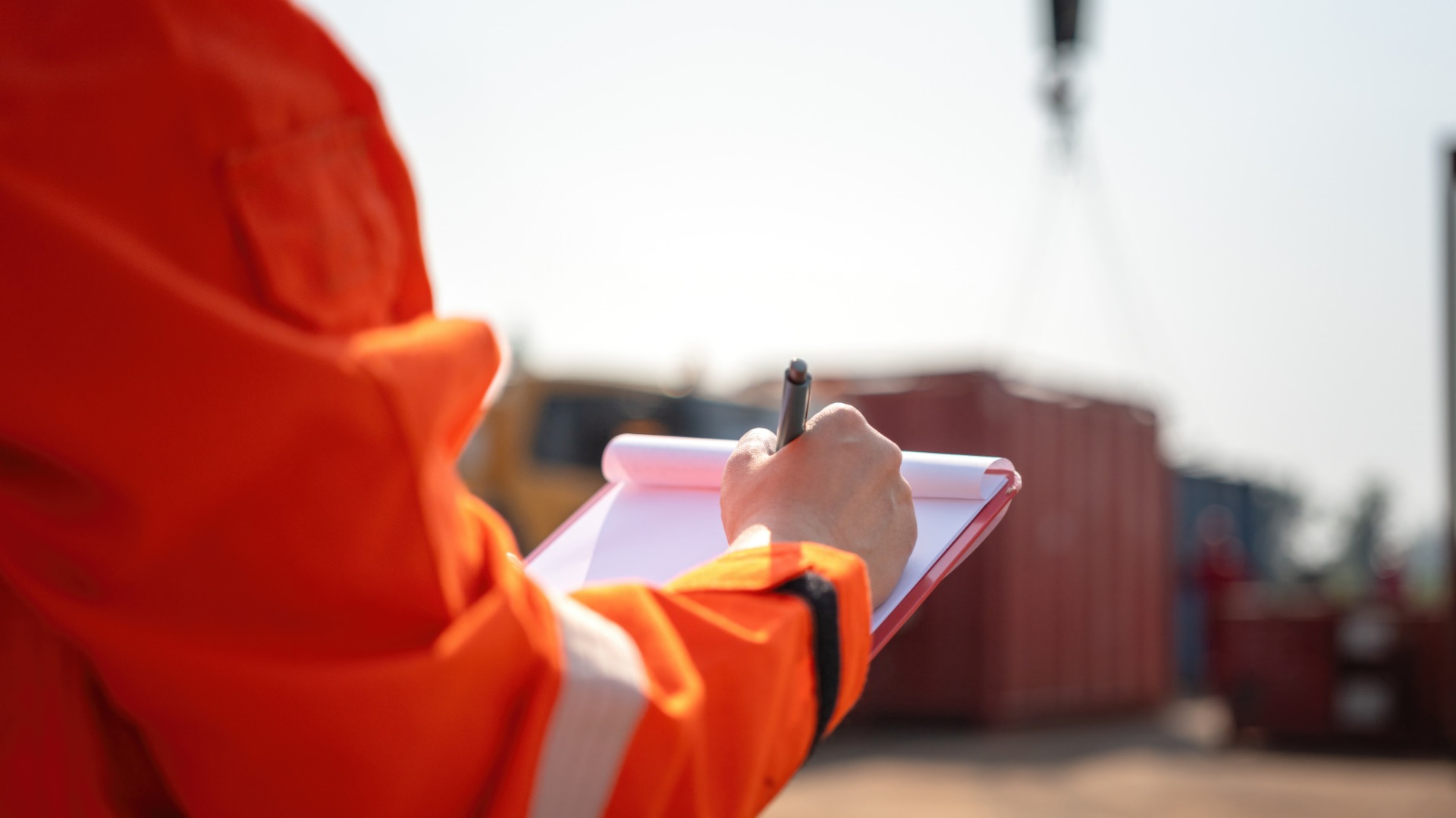 A person in an orange jacket is holding a clipboard and is marking something down on the paper that’s attached to the clipboard.