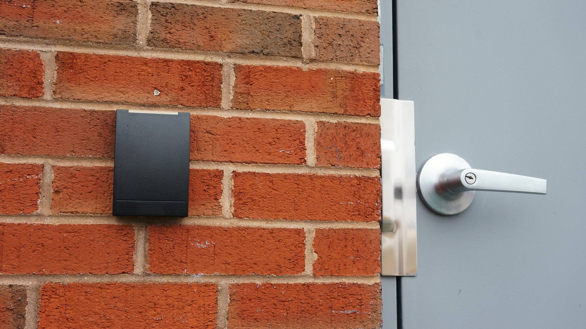 A black access control pad sits on an outer brick wall next to a light grey door with a silver handle.