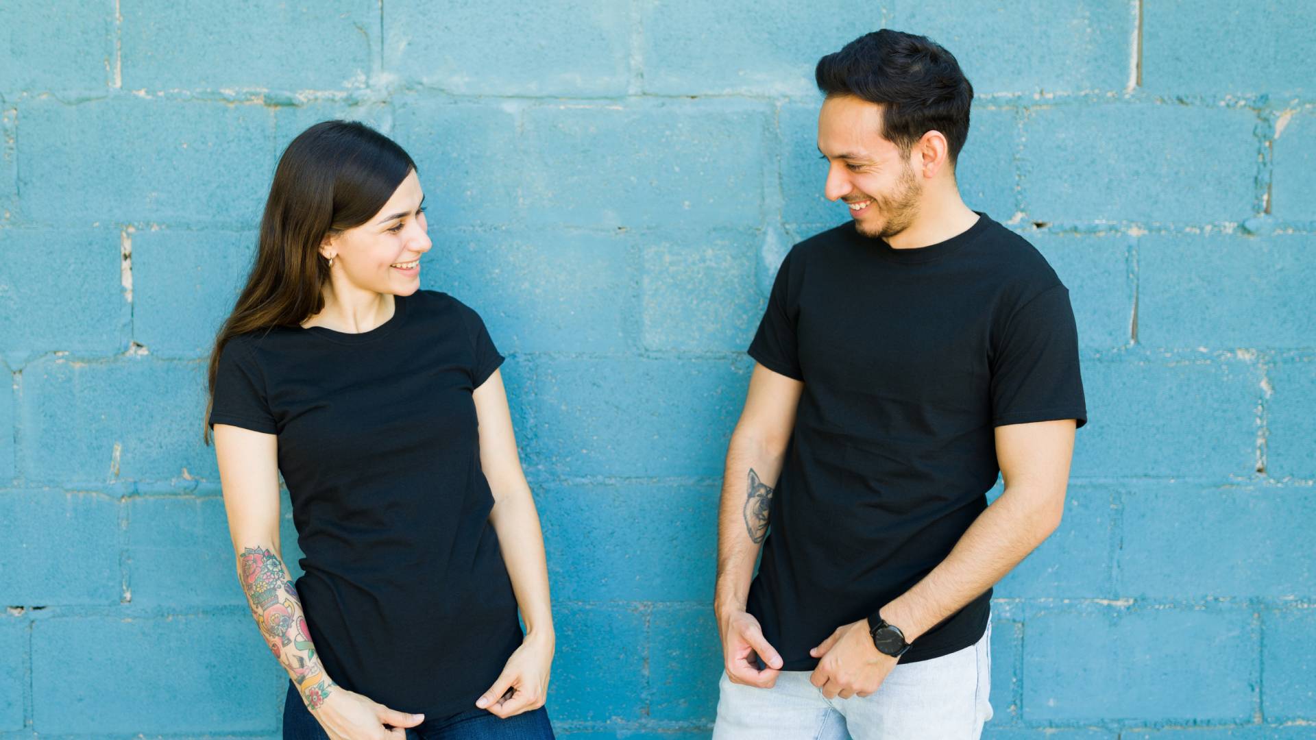 A woman and a man wearing matching black T-shirts while posing turned toward each other in front of a blue brick wall.