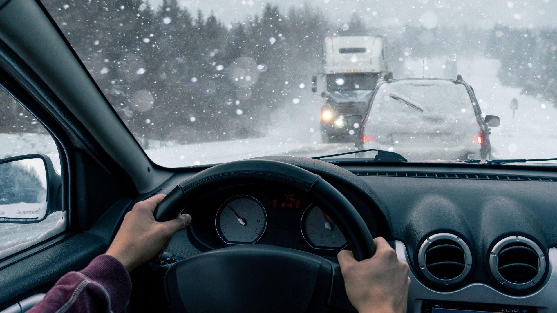 The point of view of a man driving in winter on a snowy road. Snow is falling and traffic is in front and coming toward him.