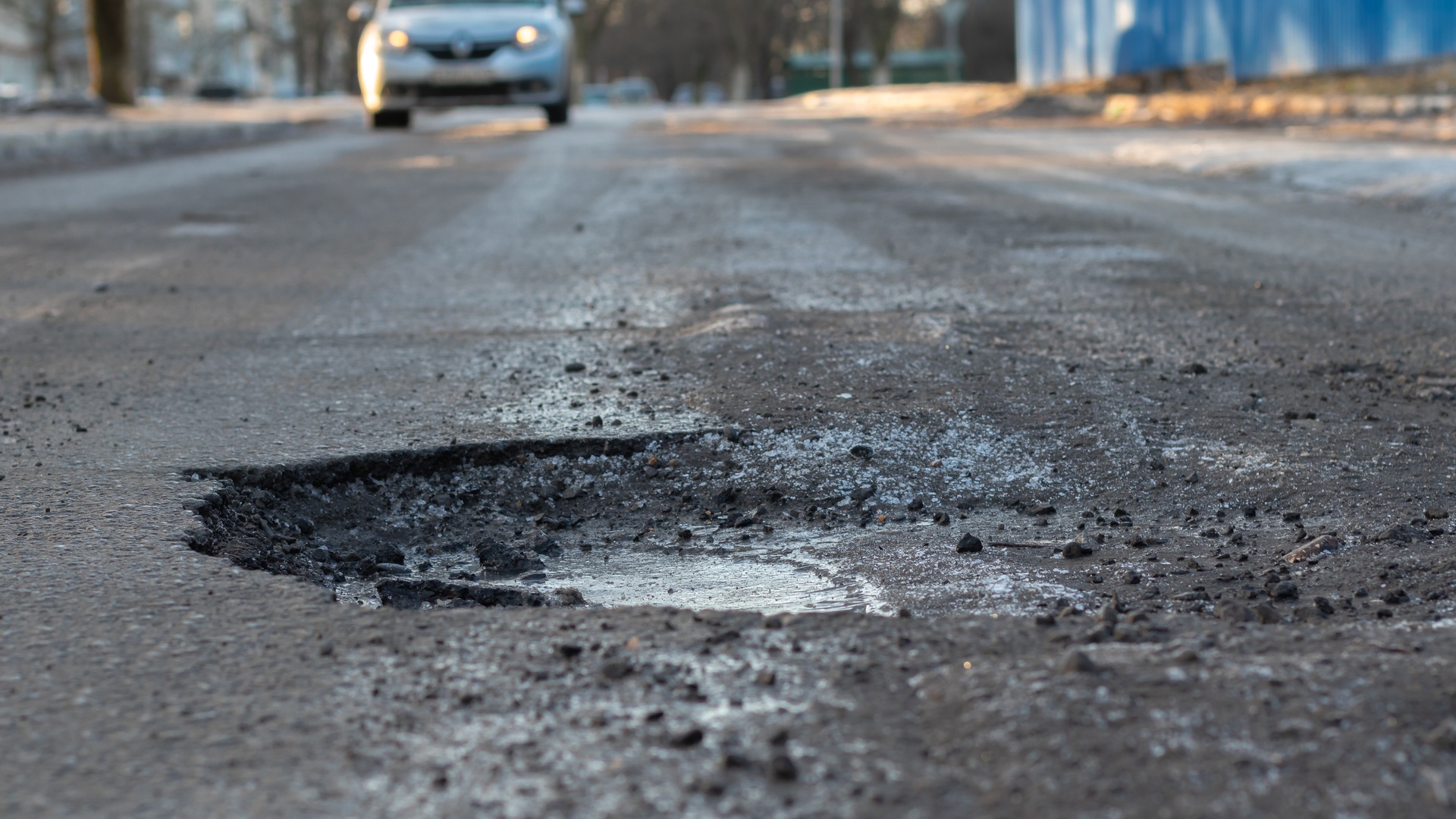 A close-up of a pothole in the middle of the road and a silver car in the background that is driving towards the pothole.