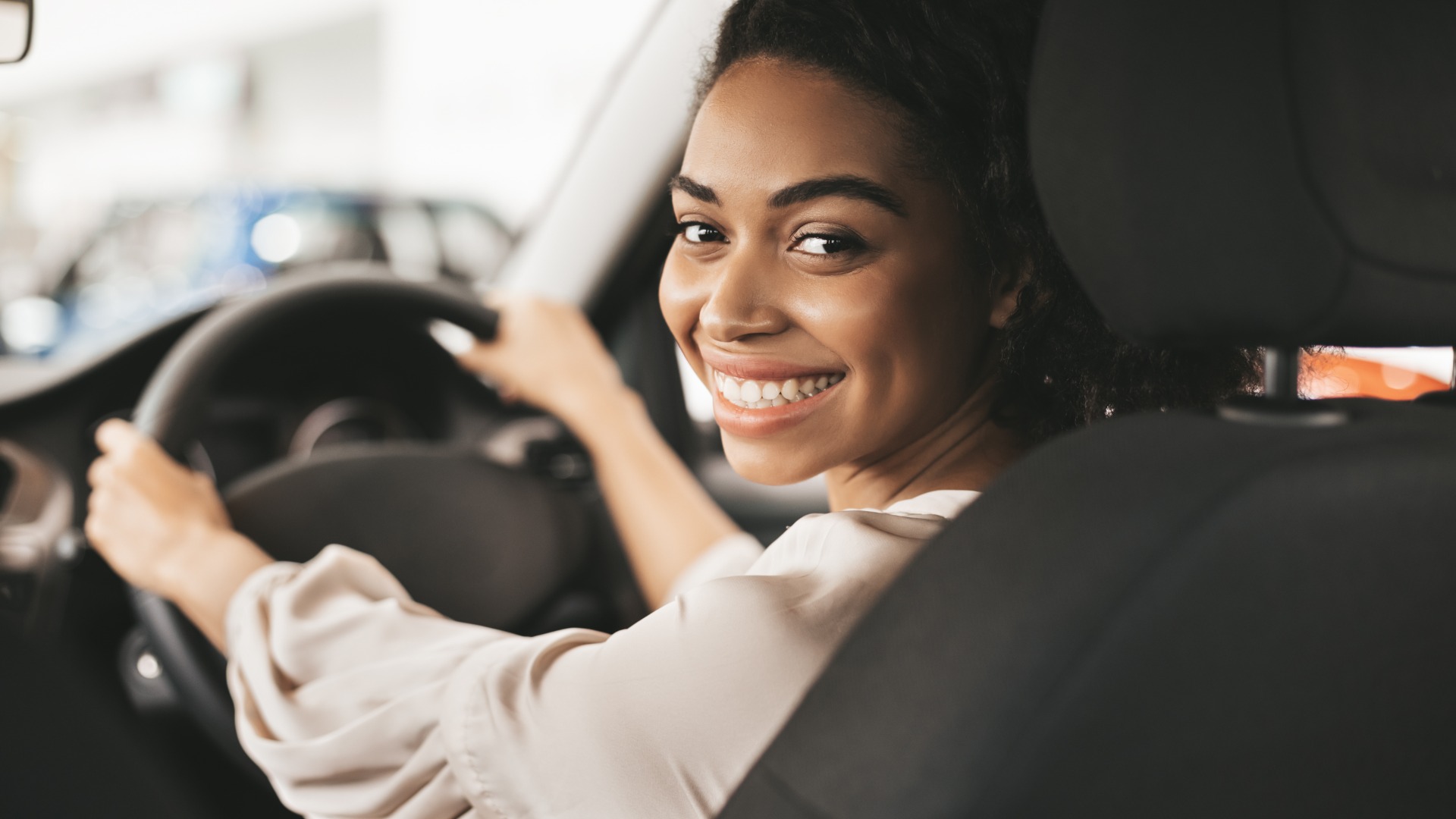 A woman sitting behind the steering wheel of a car. She is looking over her shoulder, smiling at the backseat.