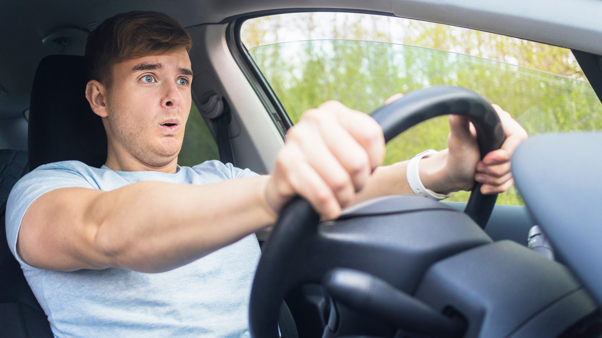 A man sitting behind the wheel of a car, looking scared. He has both hands on the wheel and is looking straight ahead.