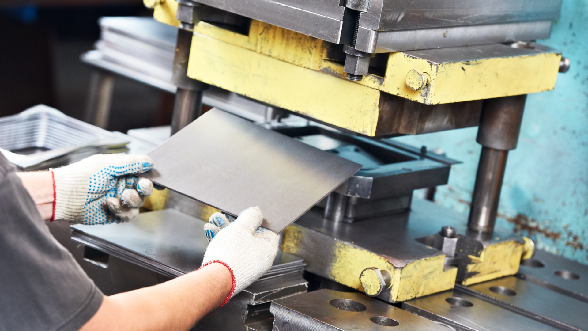 A worker wearing white gloves slides a piece of sheet metal into a metal press machine in a warehouse.