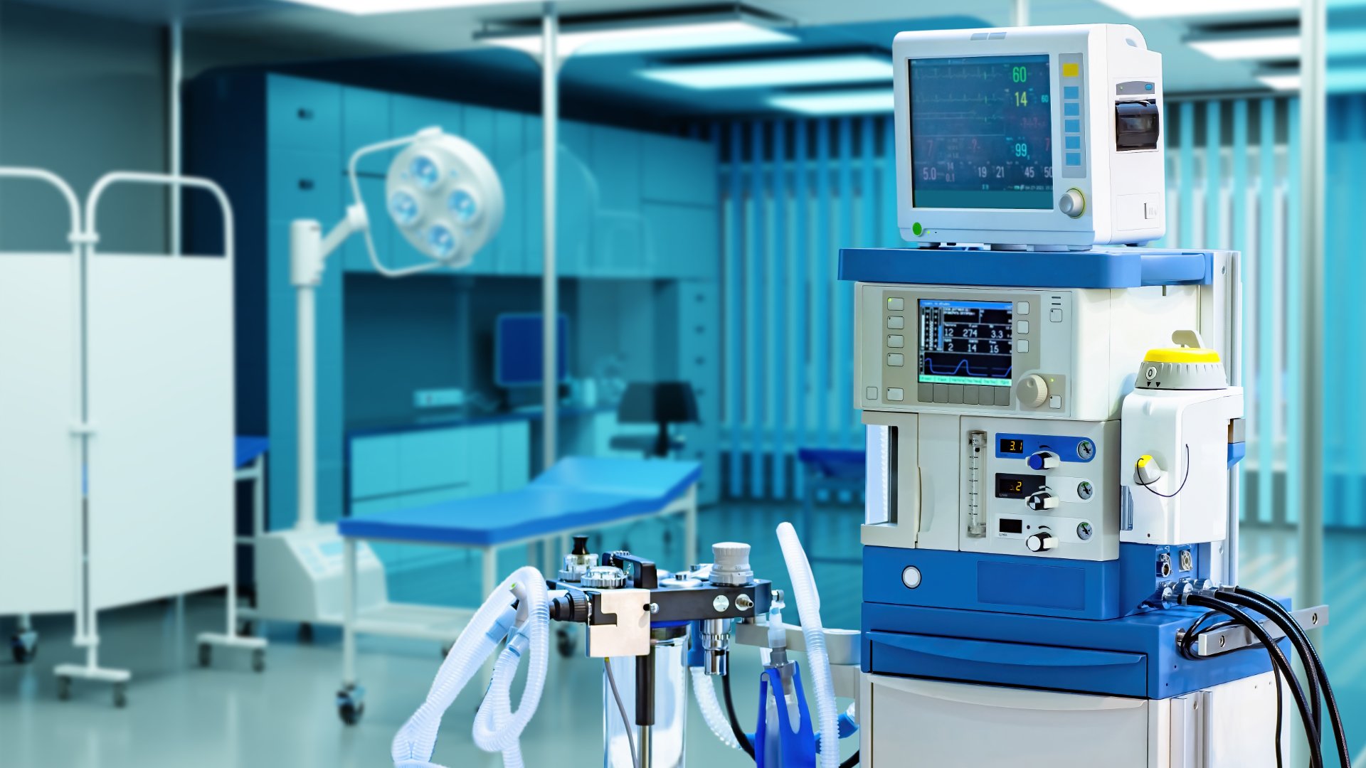 An anesthesia machine with monitors and breathing tubes standing beside an operating table in a blue medical room.