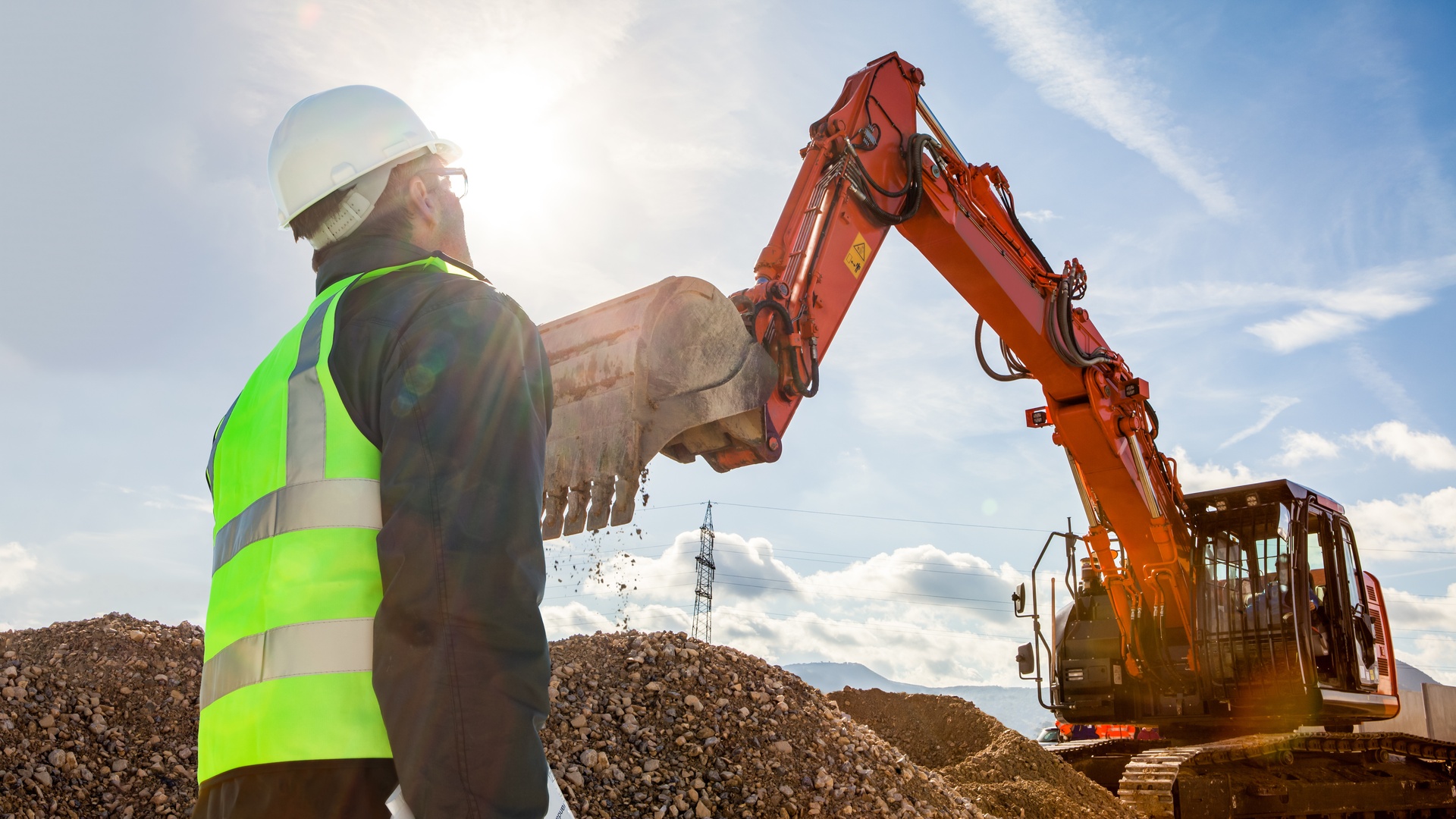 A construction worker stands on a construction site in front of a massive excavator, which prepares to pick up gravel.