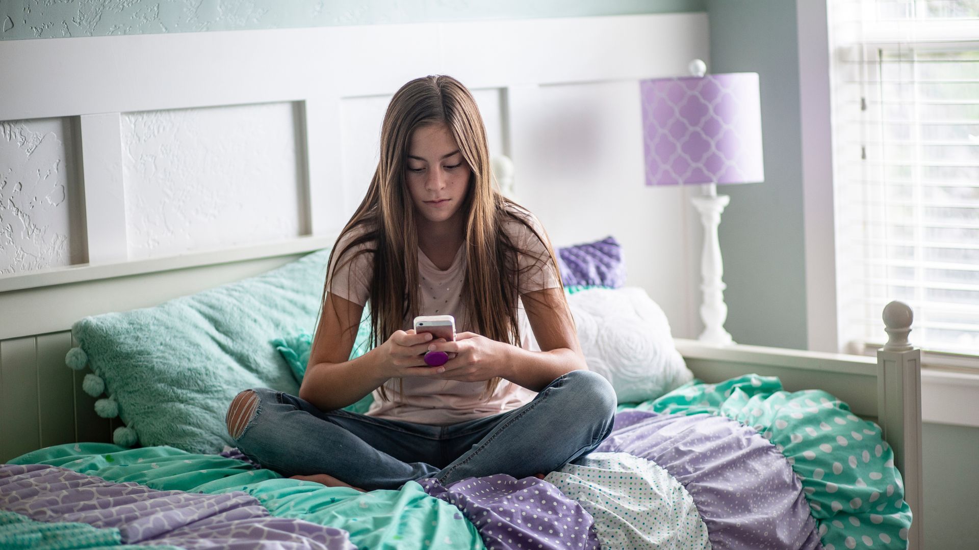 Tween girl sitting on a turquoise and lilac-purple bed in a lilac-themed room while texting on the phone.