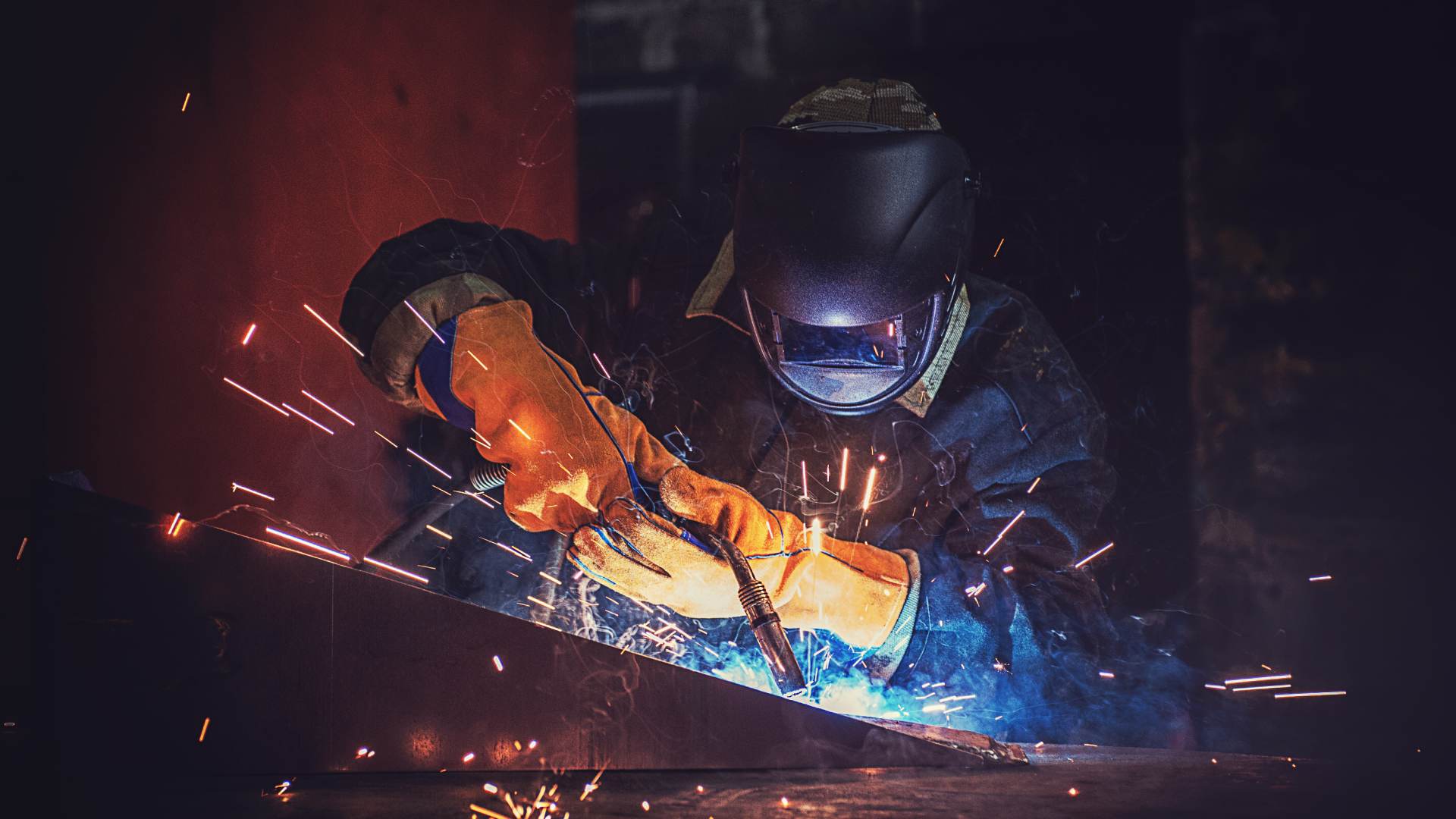 A metal fabrication worker wearing a protective mask and gloves as they hold a welding tool to weld metal in a workshop.