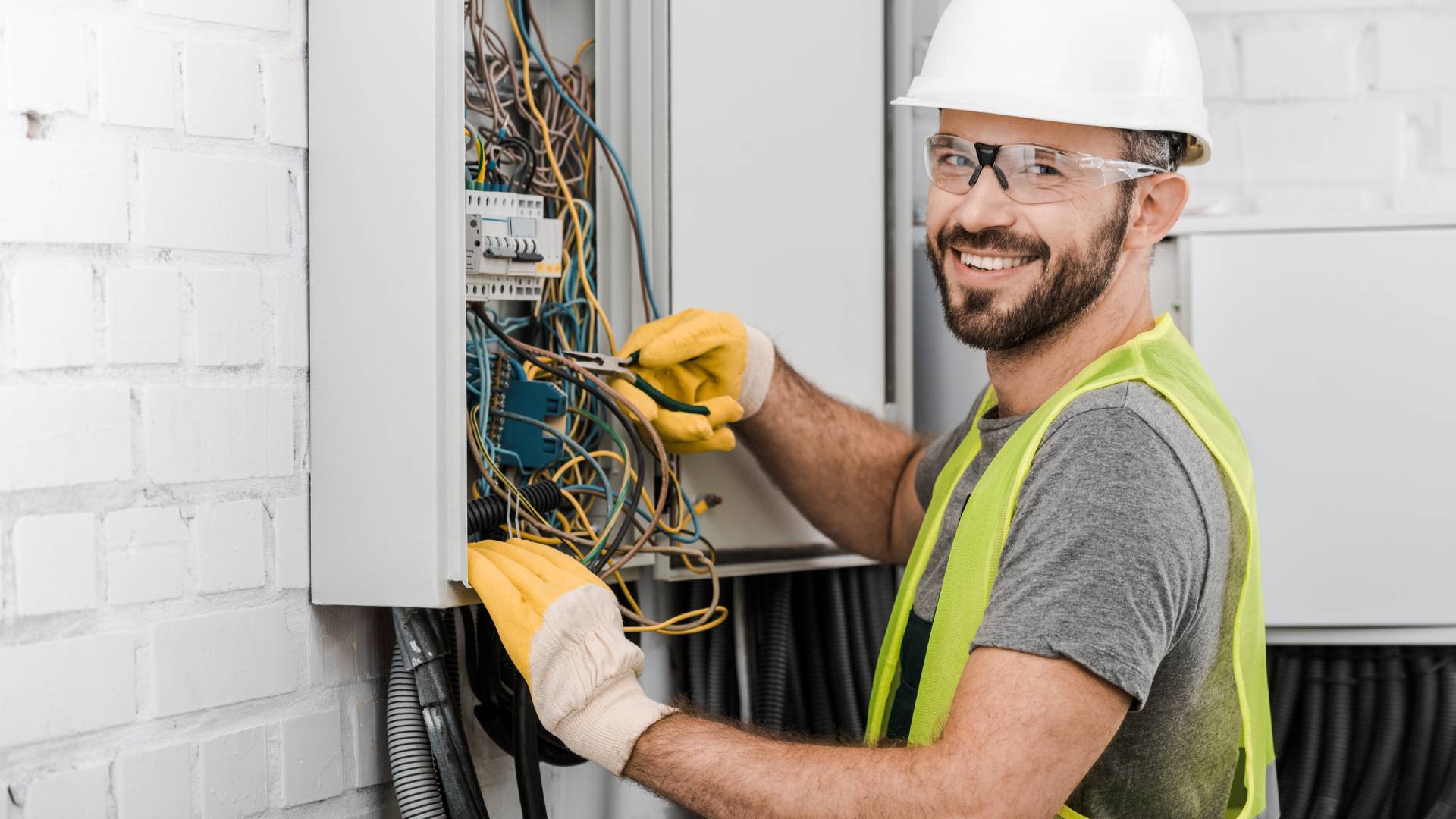 A handsome electrician smiling at the camera as he repairs an electrical box with pliers in a corridor.