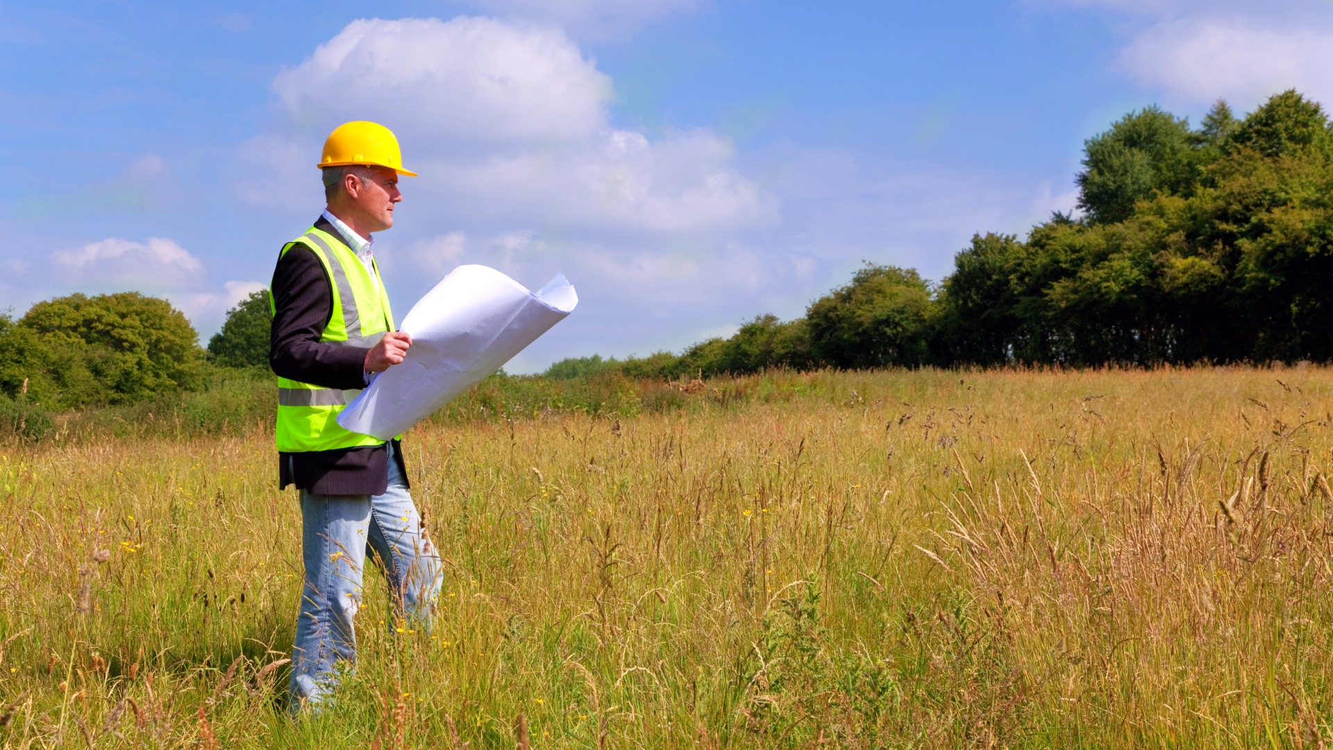 An architect holding blueprints standing in an empty land plot. There is tall grass coming up to his knees.