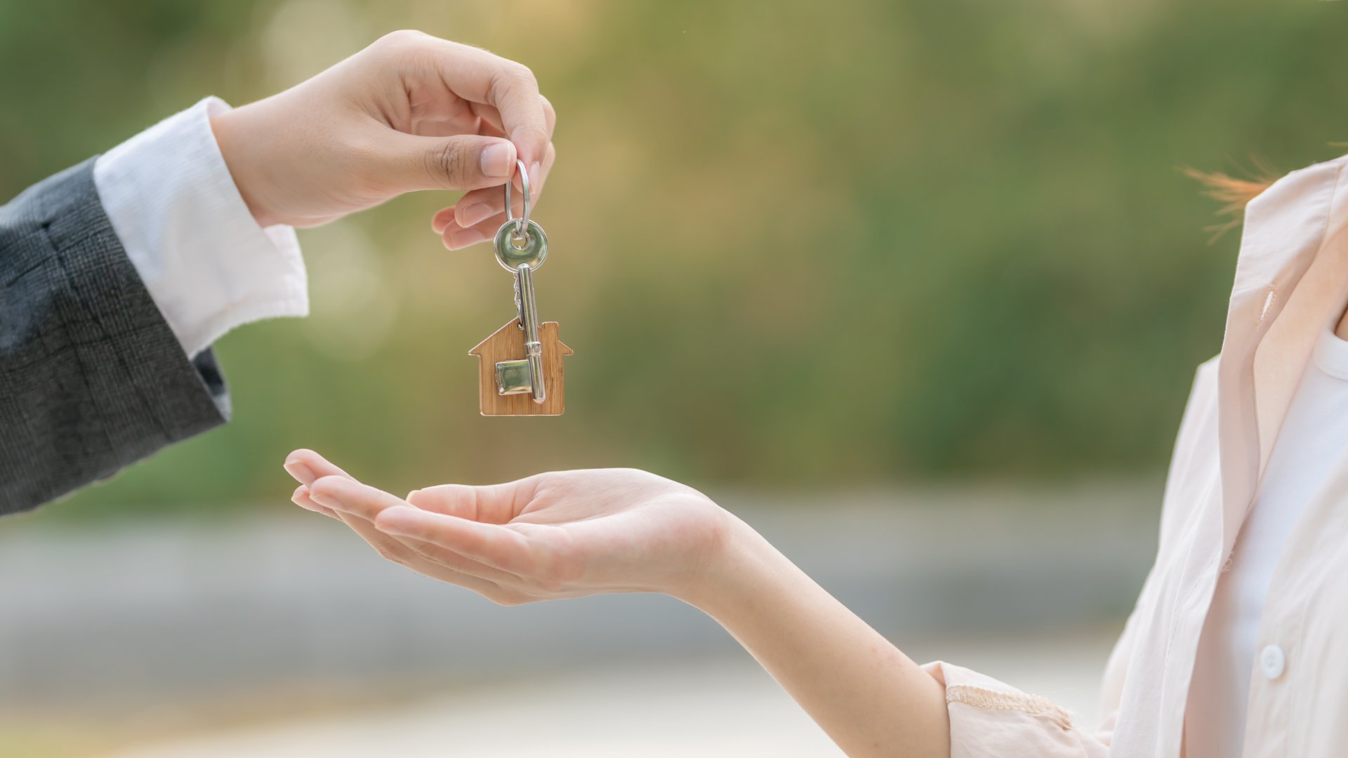 A hand in a suit jacket handing a key over to a woman with her hand outstretched. The key has a small house keychain.