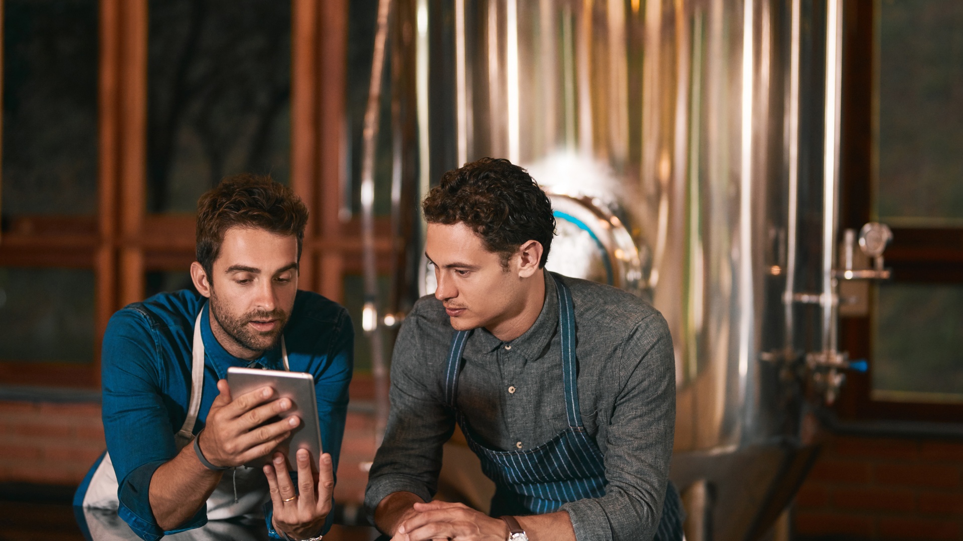Two young businessmen are in a distillery and are looking at a touch screen tablet while wearing aprons.