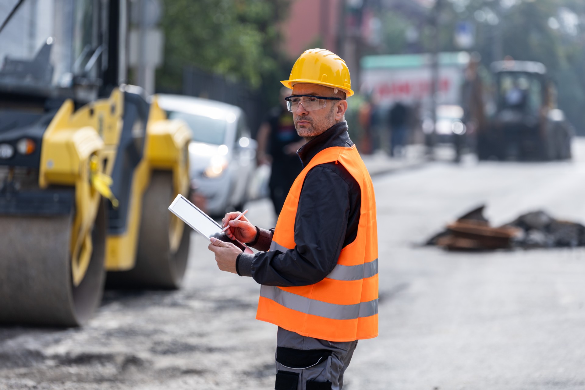 A construction worker in safety gear uses a tablet near a yellow road roller on an outdoor construction site.