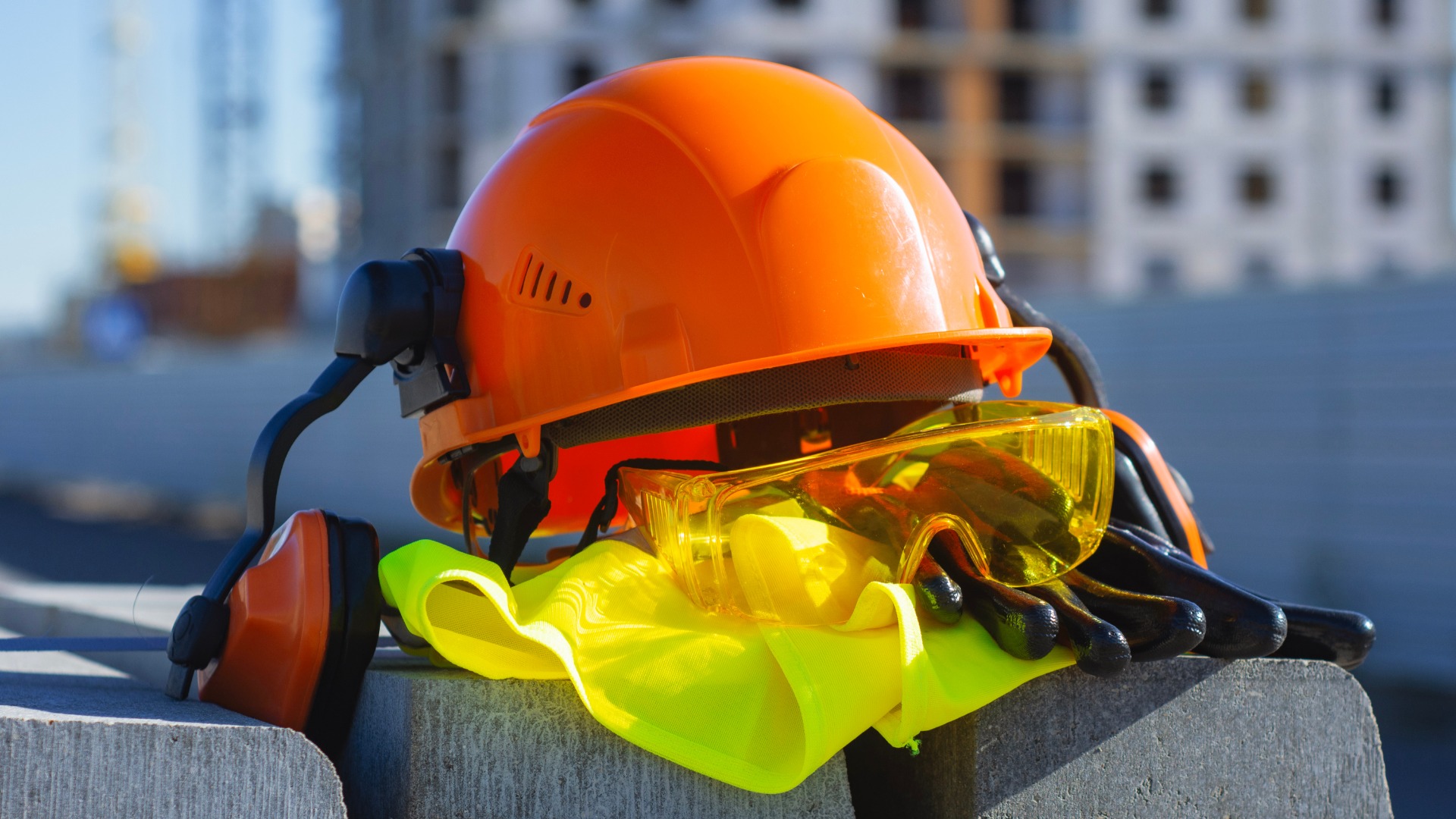 A close-up of a hard hat, earmuffs, safety glasses, gloves, and safety vest stacked neatly on top of concrete at a worksite.