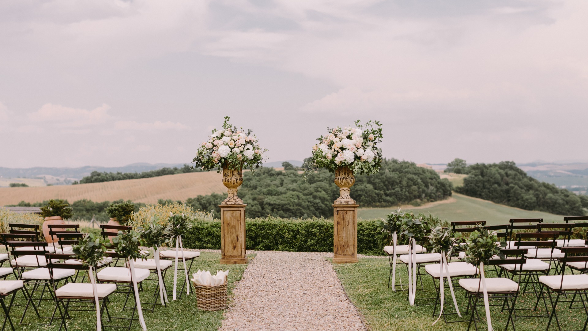 An outside aisle leads to two pedestals with large bouquets of white flowers on them. Chairs appear on either side of the aisle.