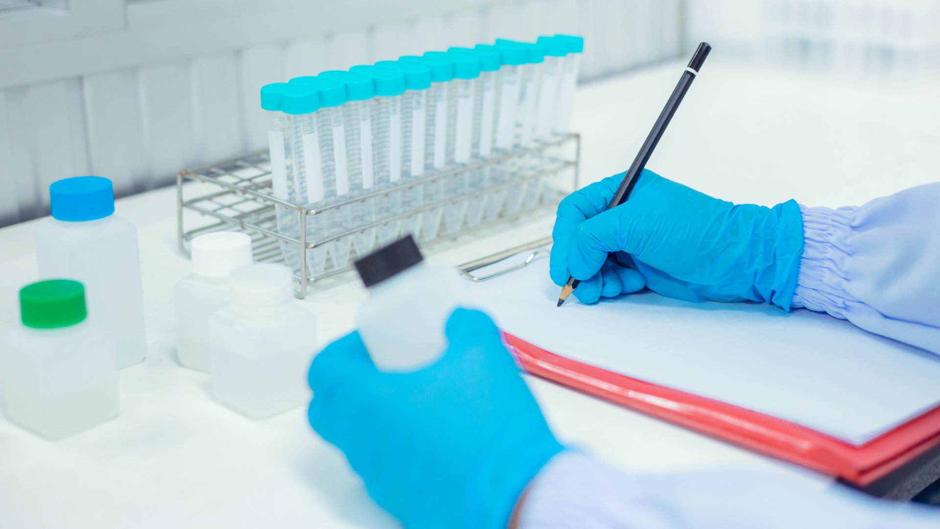 A scientist holds a plastic bottle in one hand and uses the other to write notes with a pencil on a clipboard.