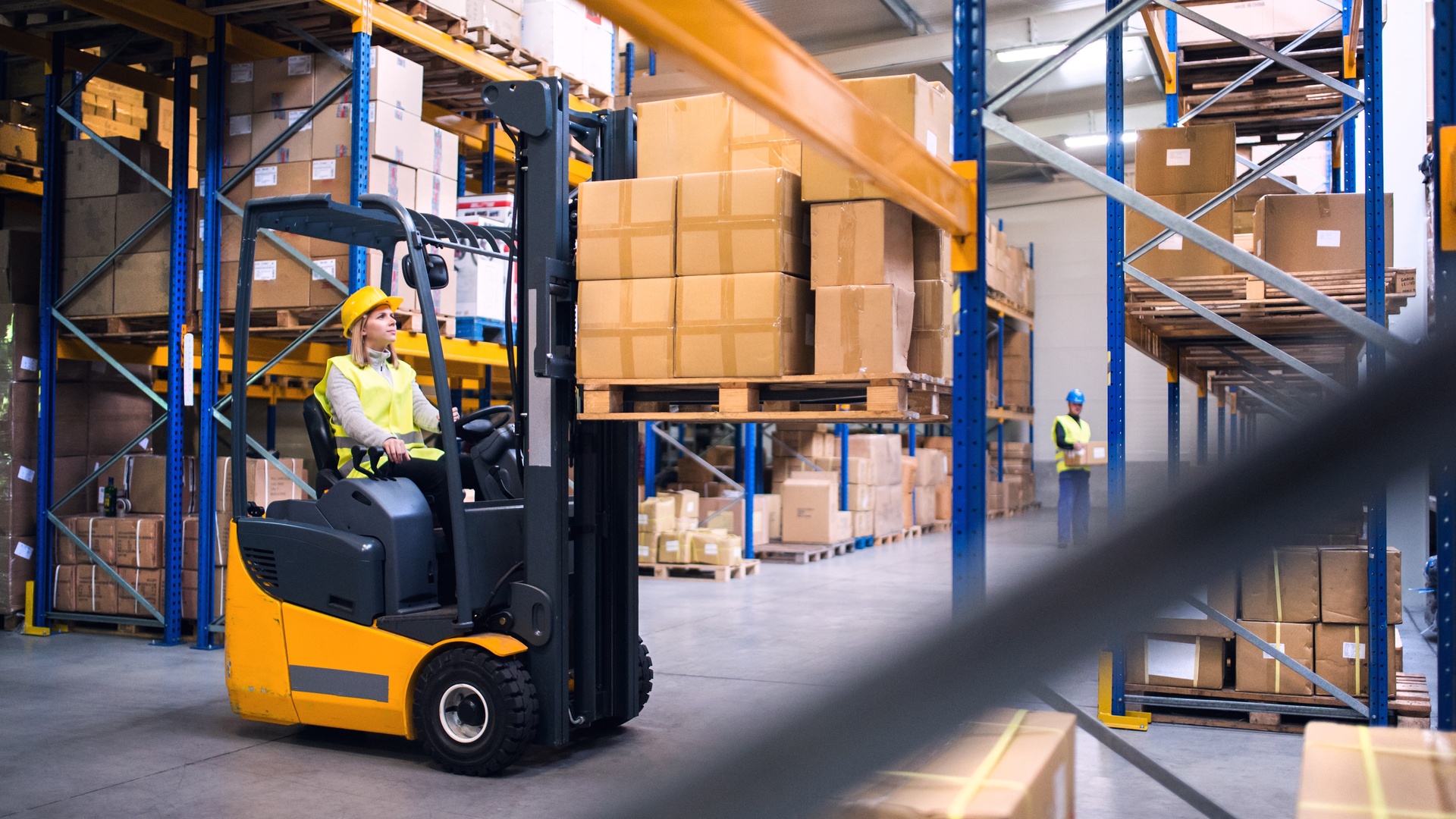 A woman in a yellow hard hat and safety vest operates a forklift, moving stacks of cardboard boxes in a warehouse.