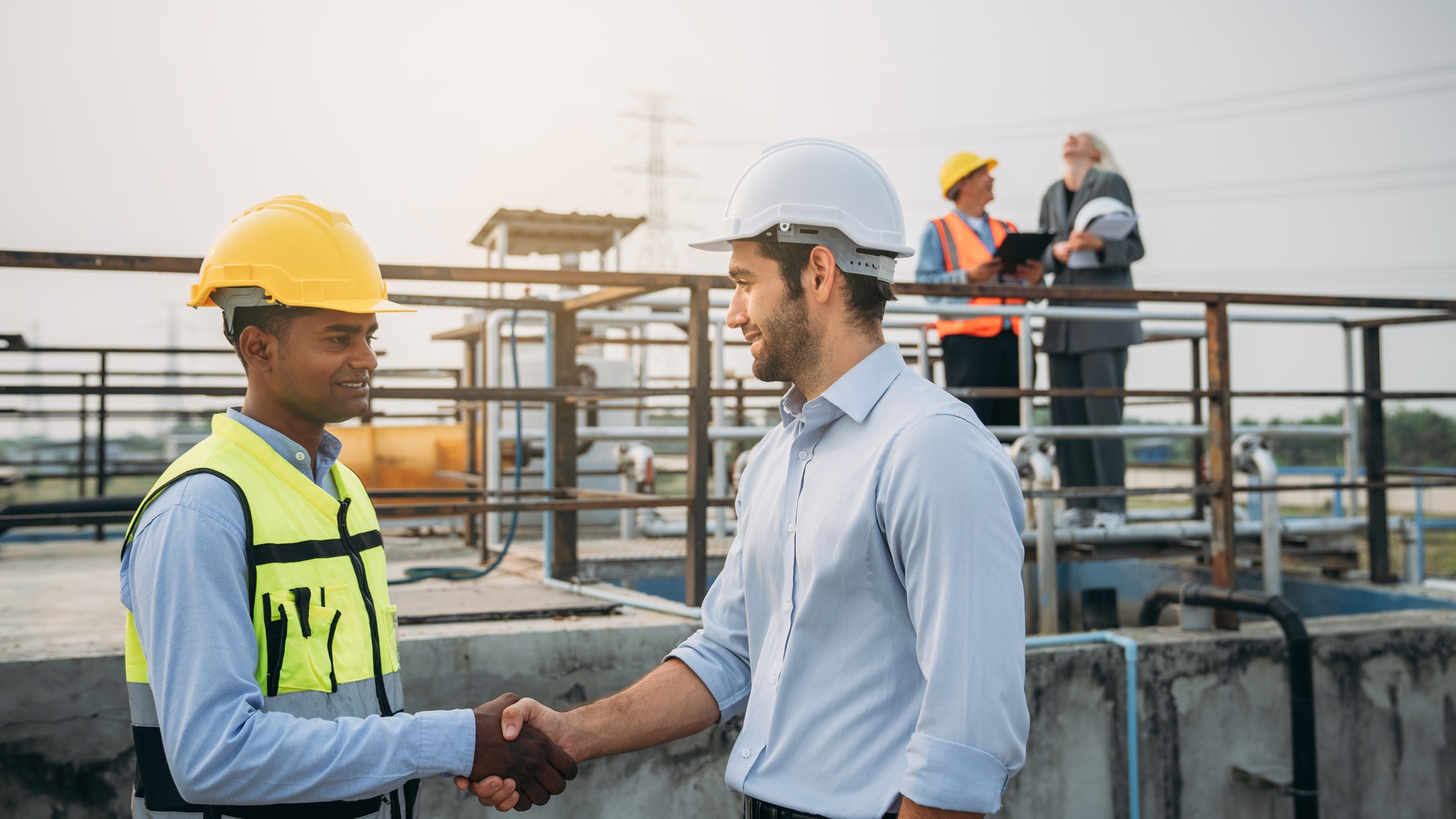Two workers in hard hats shaking hands at an industrial site, with equipment, pipes, and other workers in the background.