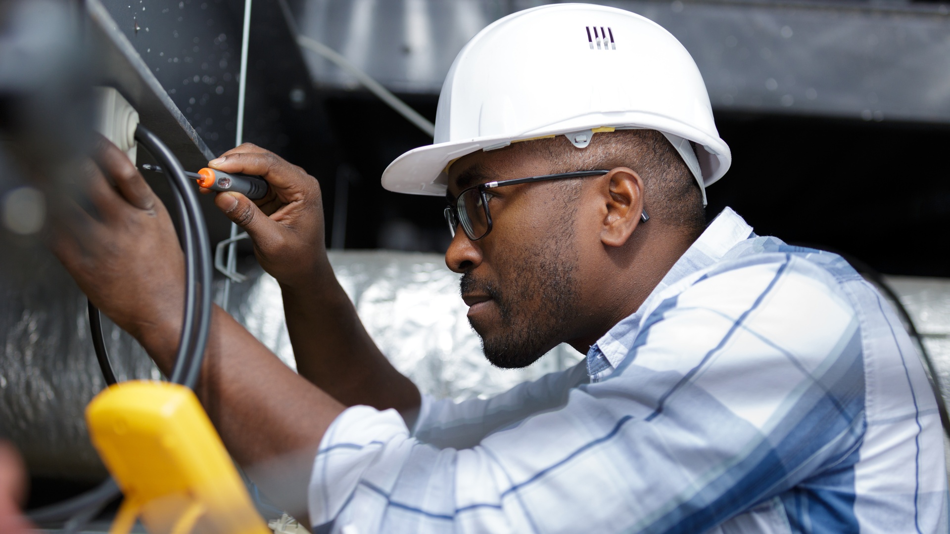 An electrician is fixing cables. He's wearing a white hard hat and glasses and is concentrating on his task.