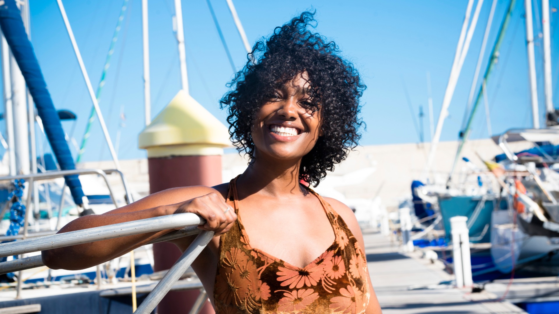 Smiling woman with curly hair stands on a marina dock, holding a railing with sailboats moored in the background.