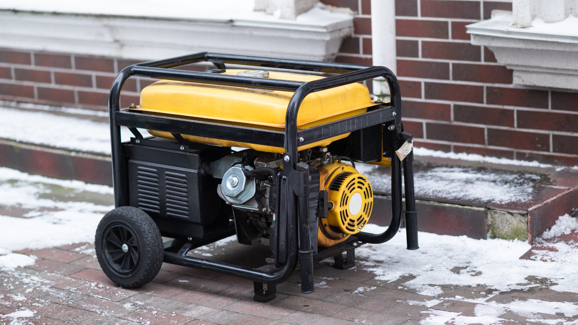 A yellow and black portable gasoline generator with two wheels outside on a brick surface covered with snow.