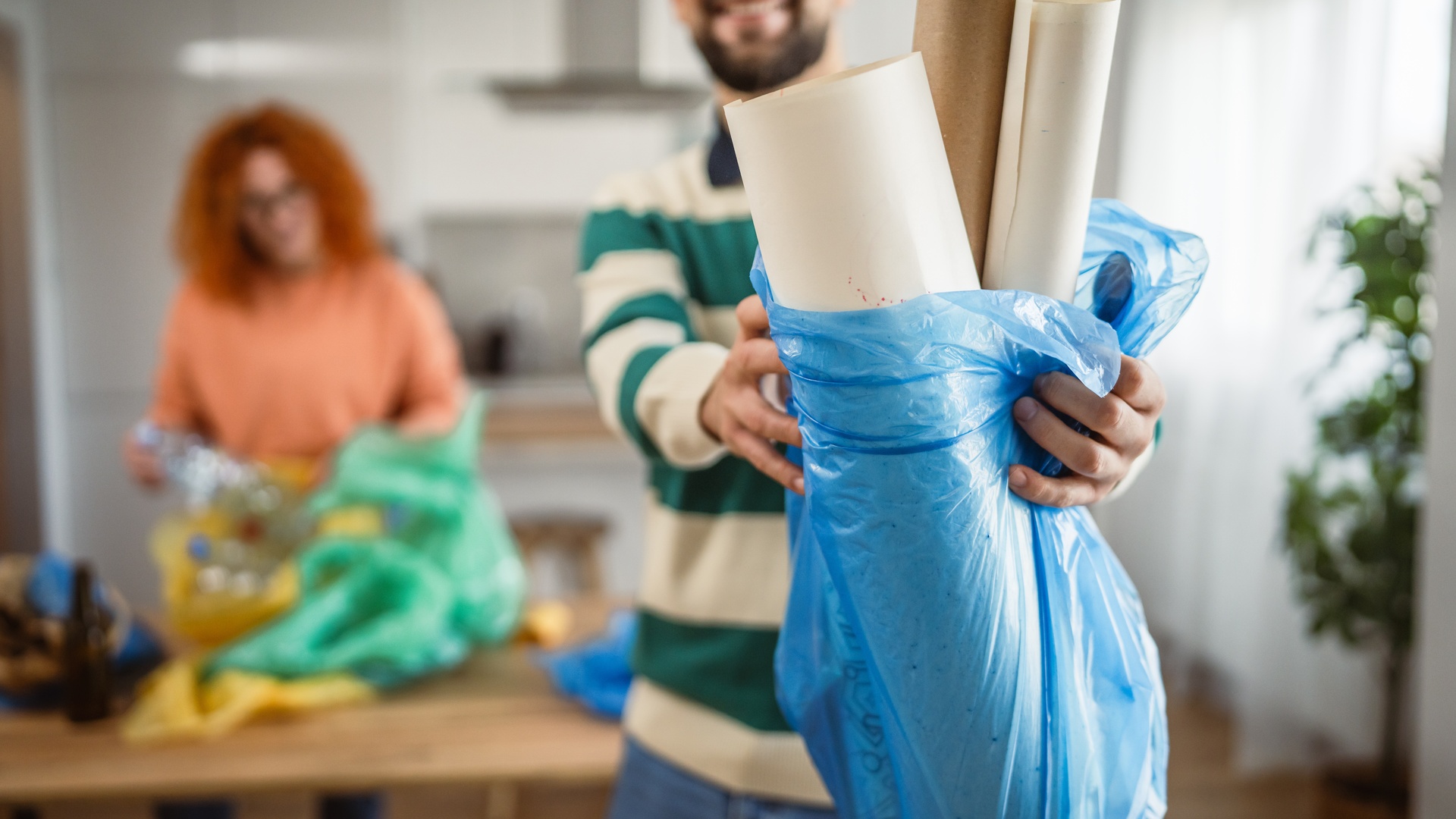 A man in a green striped sweater and brown glasses is throwing poster paper in a blue garbage bag at home.