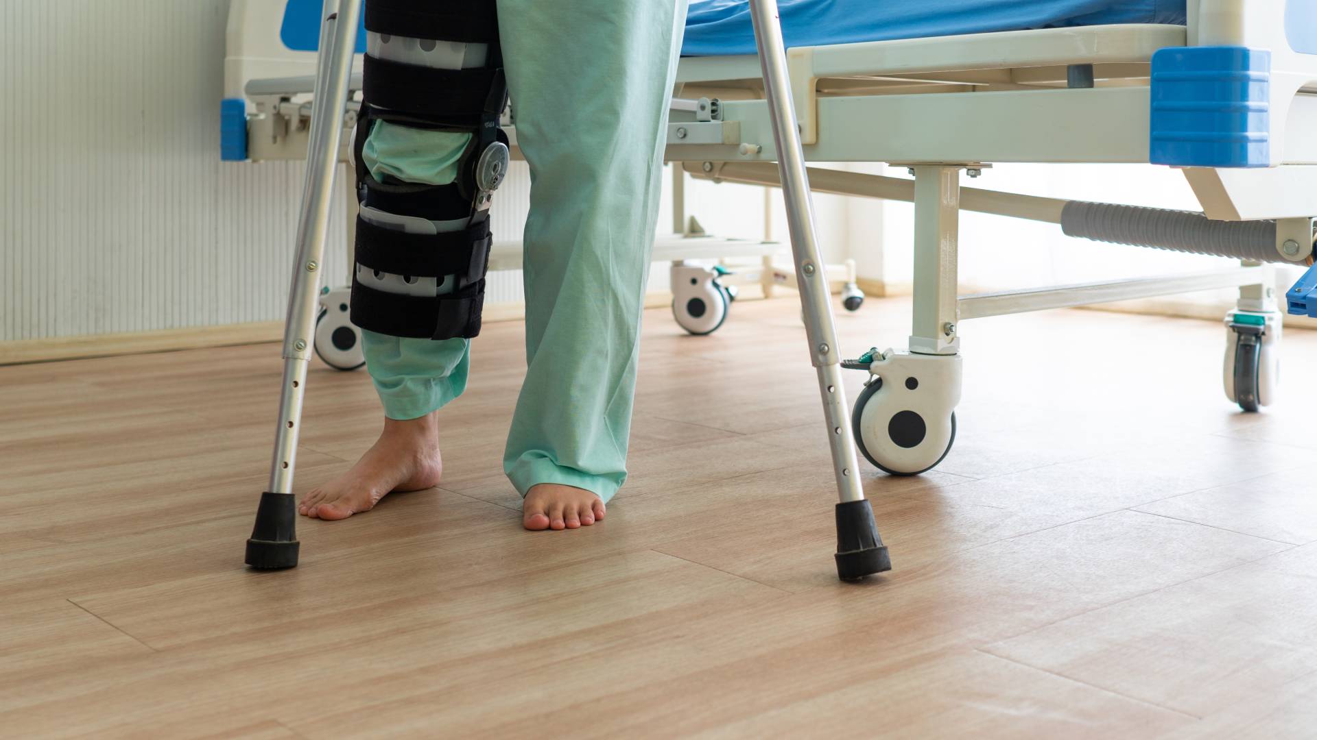 View from the thighs down of a patient wearing scrubs and a leg brace. The patient uses crutches to stand by a hospital bed.