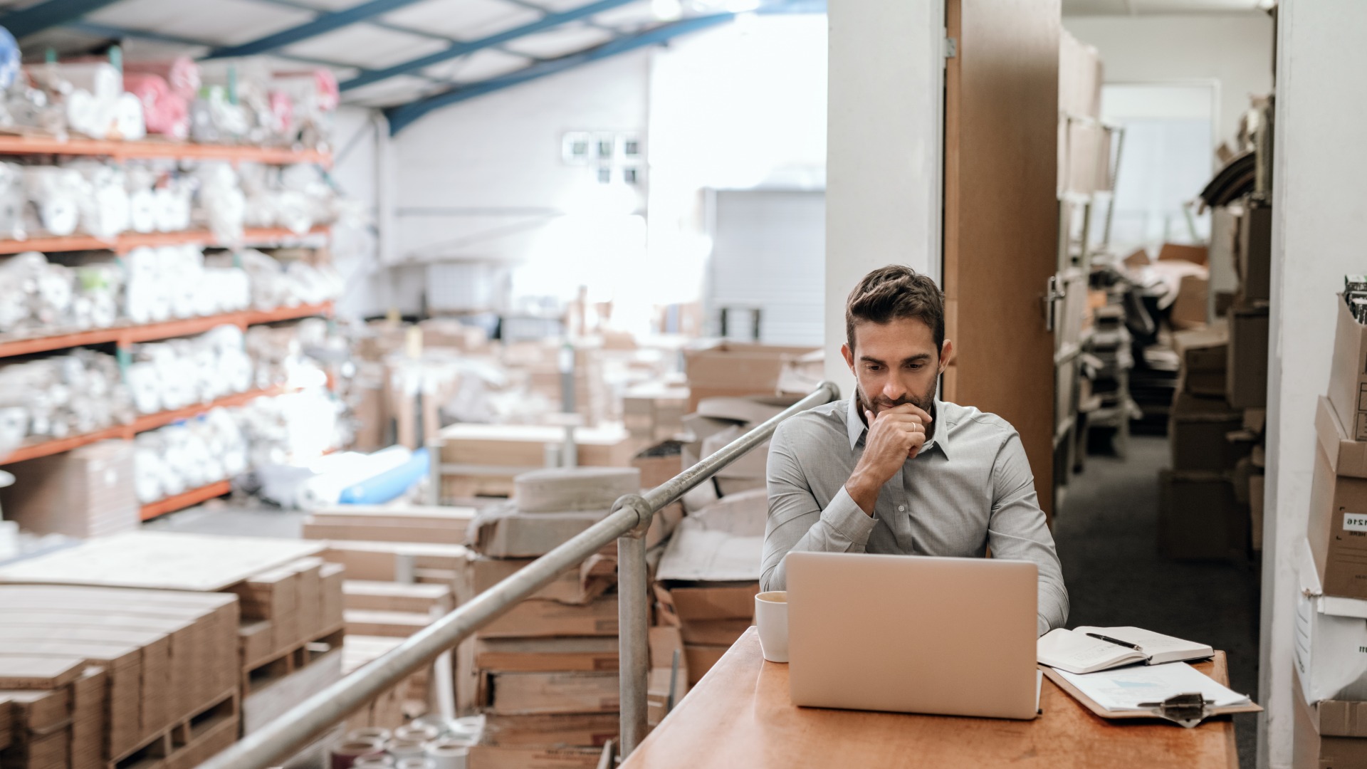 A man sitting at a desk near an office overlooking a full warehouse floor. He is looking at a laptop on the desk.