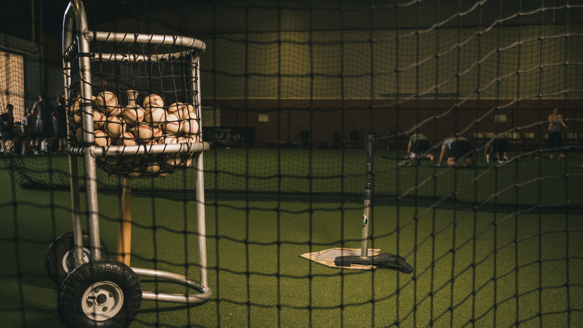 Looking through the netting of a baseball batting cage with a home plate, a tee, and a basket with baseballs.