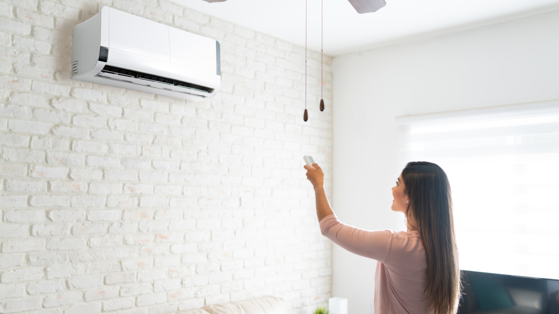 A woman with brown hair in a pink shirt uses a remote to operate a mini split mounted on a white brick wall.