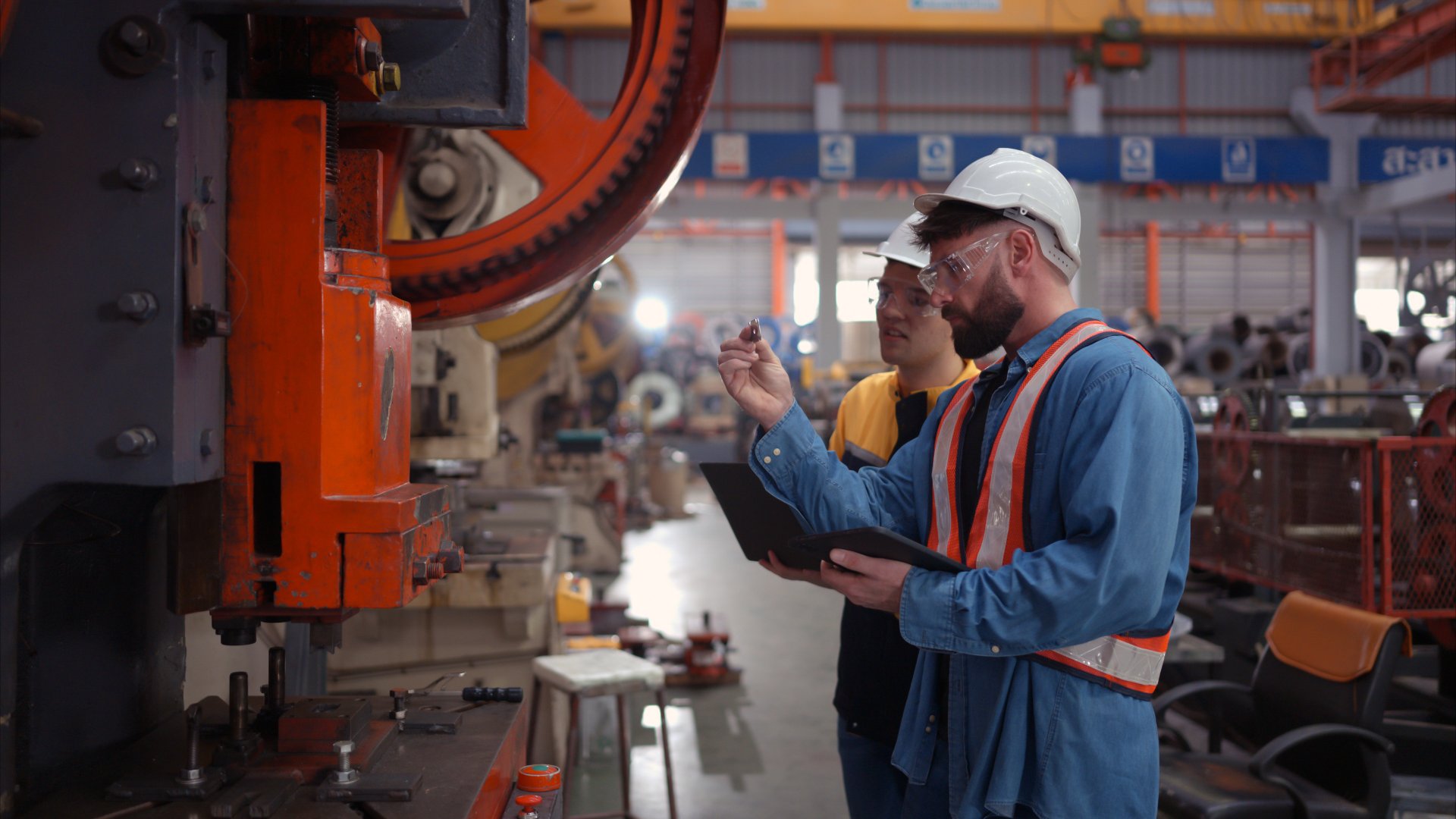 Two factory workers with hard hats and safety vests inspect a machine. One holds a clipboard, while the other has a tablet.