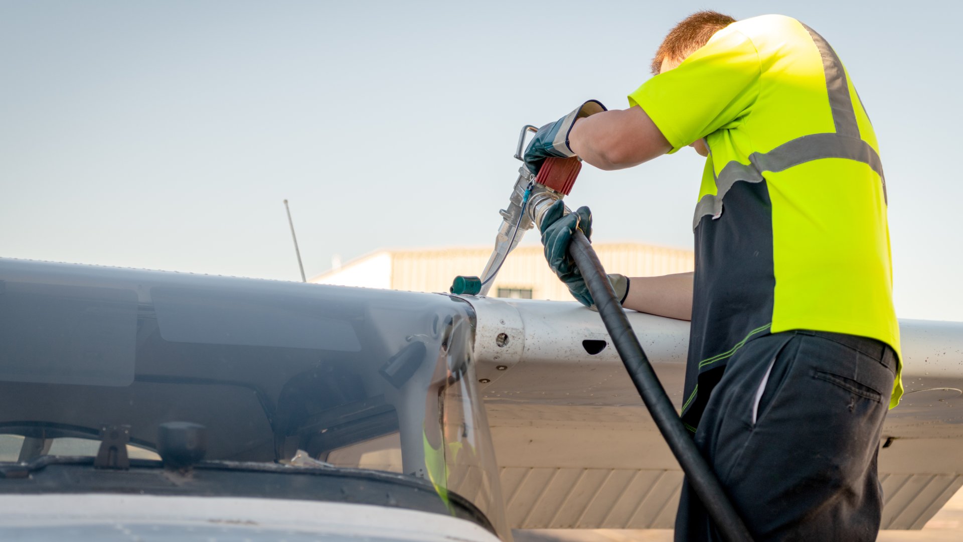 A mechanic fills up a plane's tank with fuel. The mechanic wears blue gloves and a yellow and black shirt.