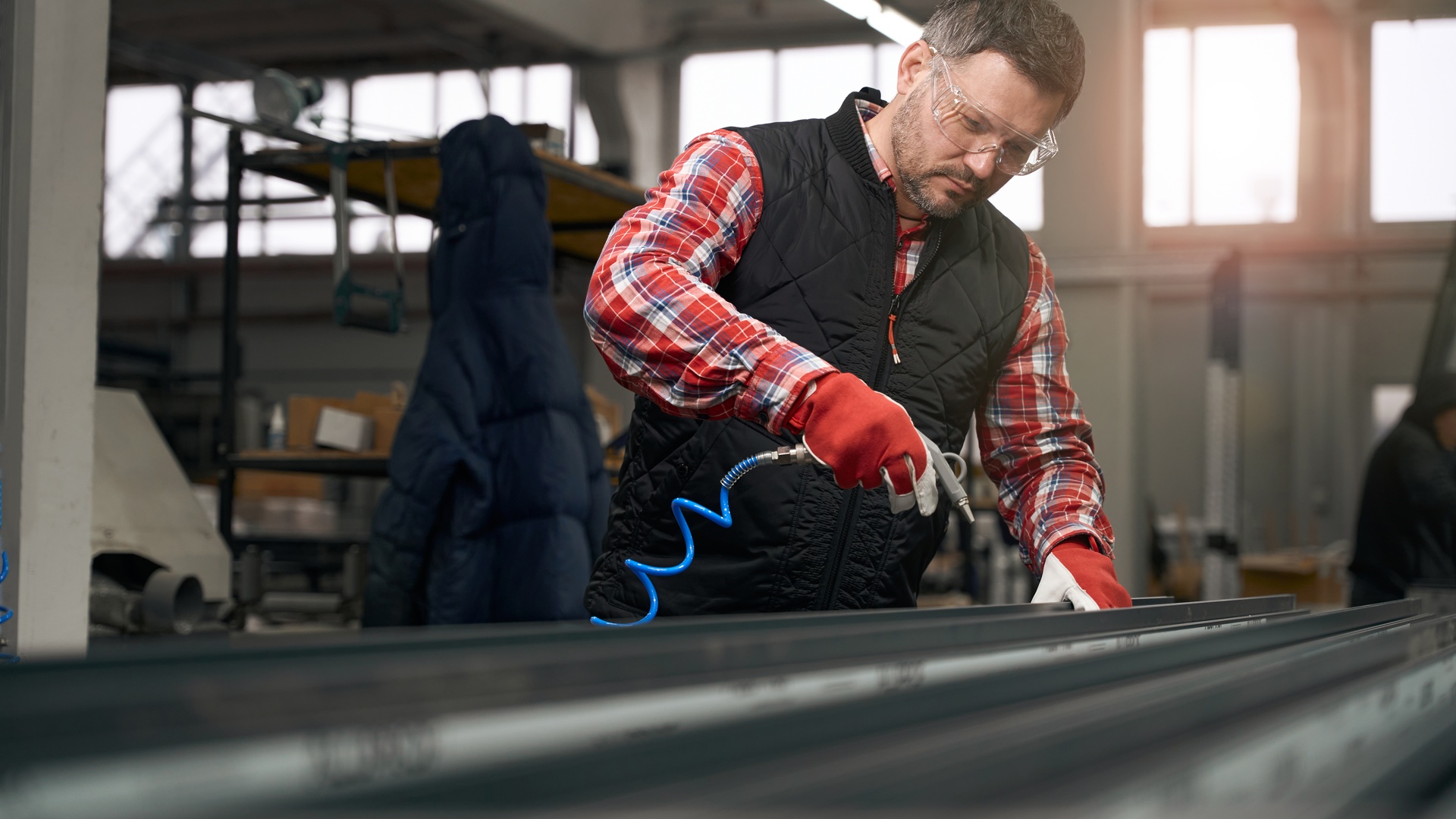 A man in a plaid shirt, black vest, safety glasses, and gloves uses a tool with a compressed air line.