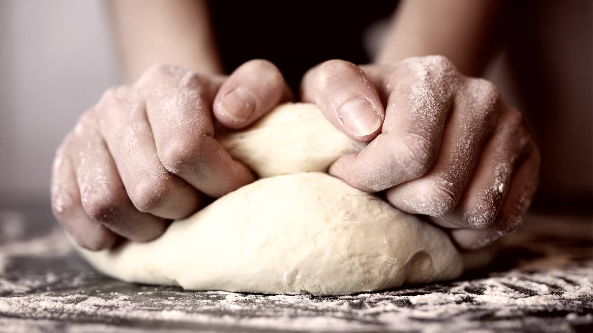 An extreme close-up view shows two flour-covered hands kneading dough on a flour-covered dark surface.