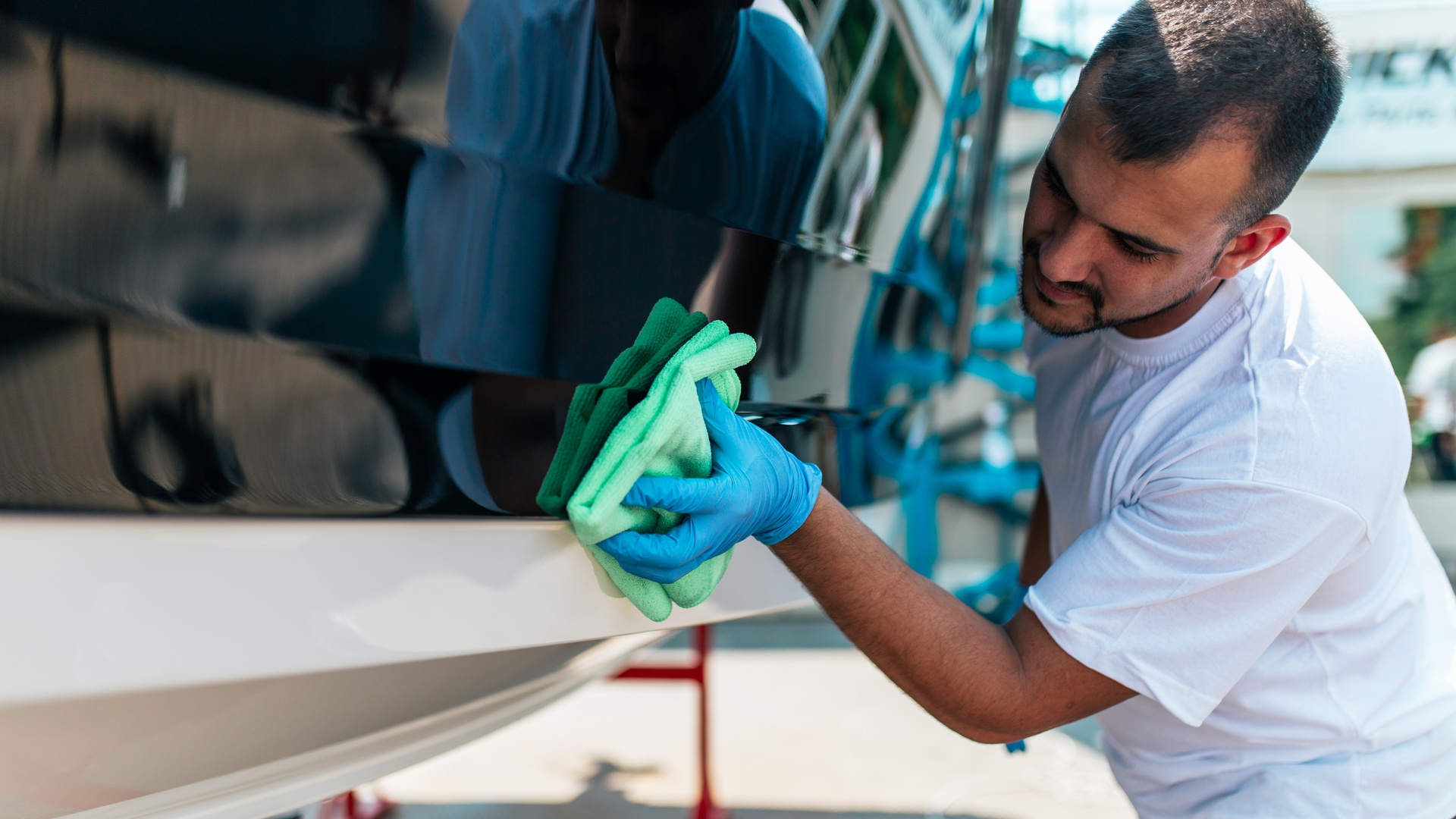A man is outdoors on a sunny day, cleaning and polishing his boat with a light blue-green microfiber cloth.