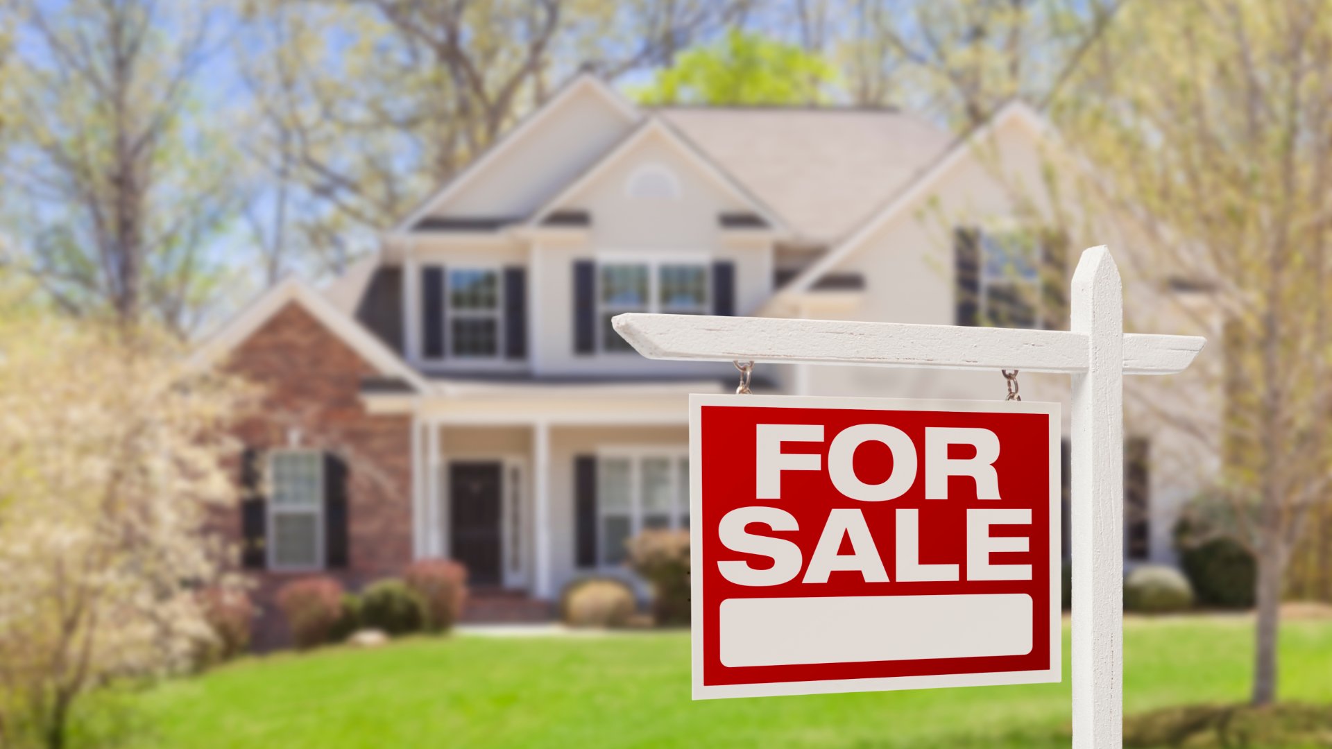 Blurry in the background is a two-story home, and in the foreground is a red for sale sign.